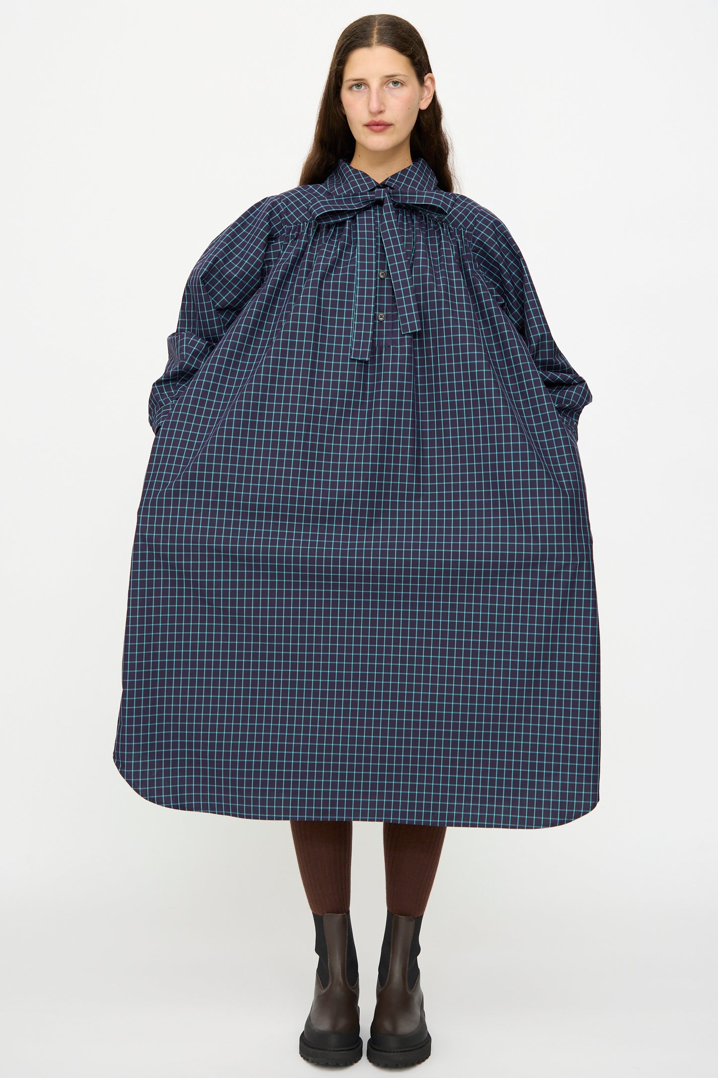 A woman stands against a plain background wearing the KasMaria Cotton Poplin Smock Dress in Blue Check, paired with brown tights and black boots.