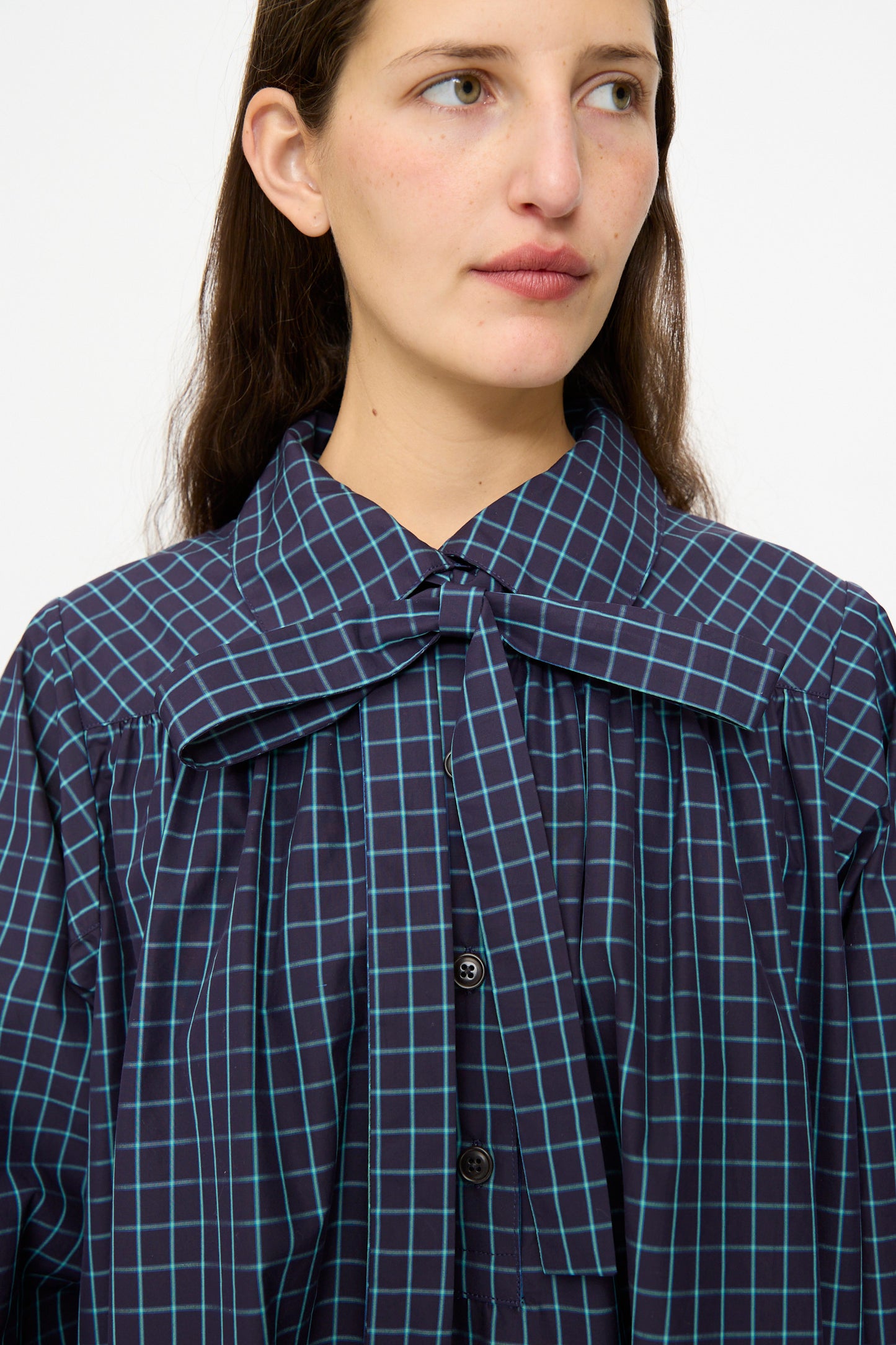 A person wearing the KasMaria Cotton Poplin Smock Dress in blue check, featuring a large bow collar, facing slightly to the right.