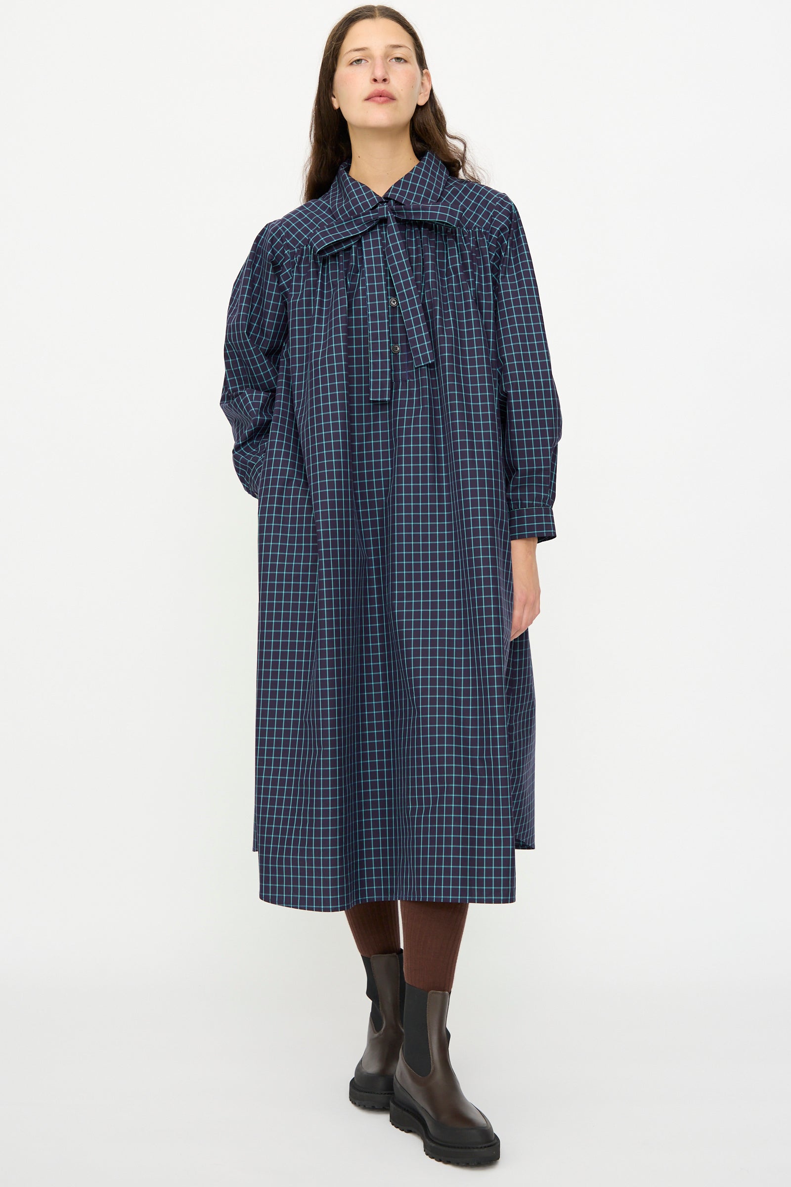 A person wears the KasMaria Cotton Poplin Smock Dress in Blue Check, styled with brown leggings and black ankle boots, standing against a plain white background.