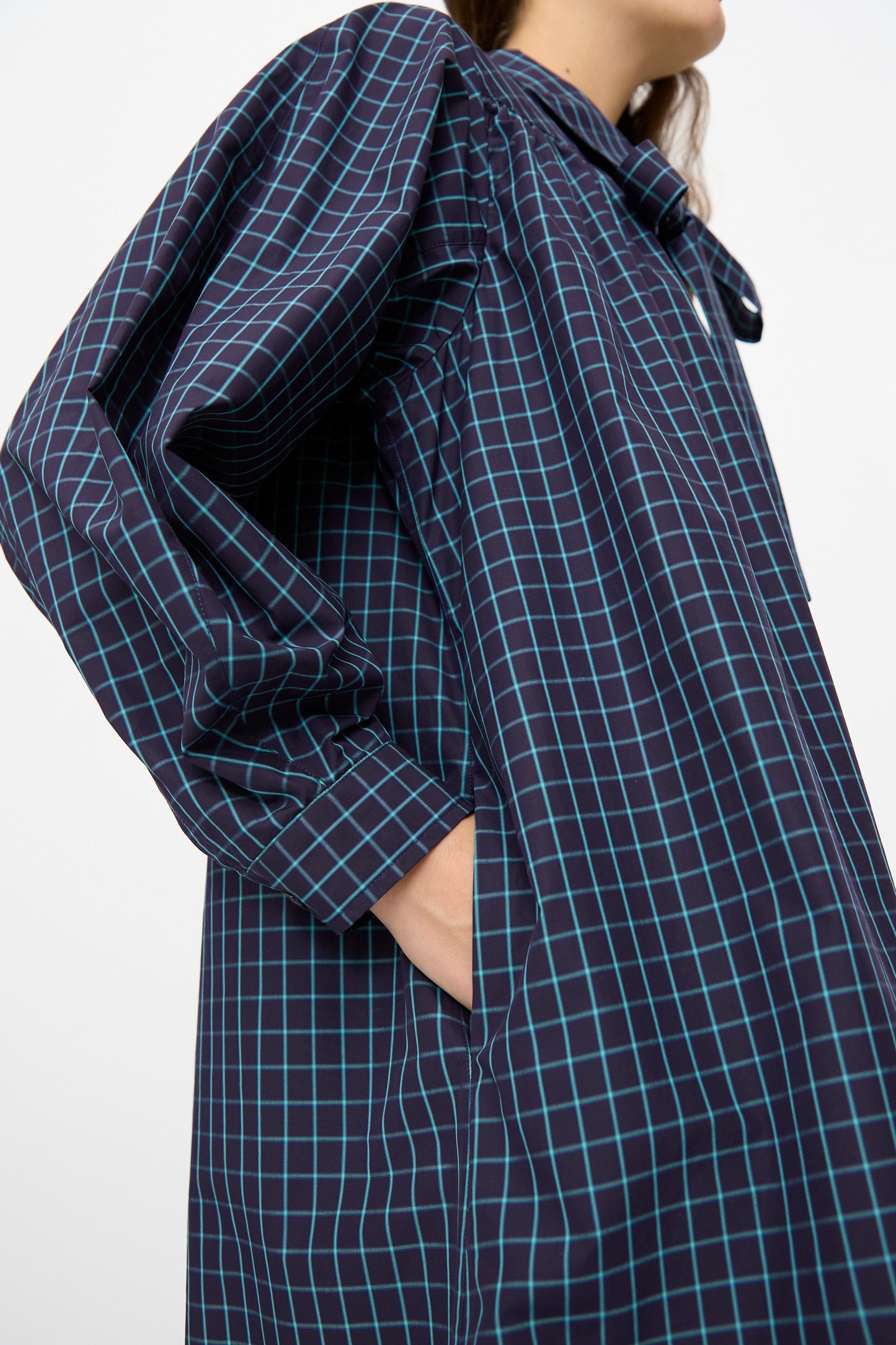 A person wears the KasMaria Cotton Poplin Smock Dress in Blue Check—an oversized navy Italian cotton poplin dress with a light blue check grid, standing with one hand in a side pocket.