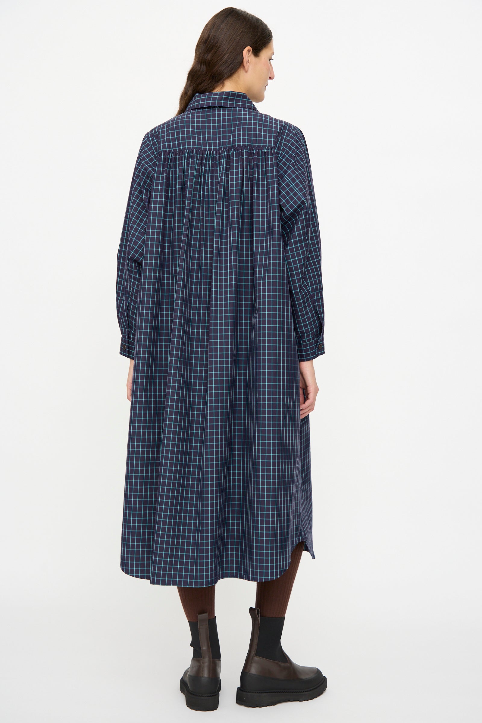 A person faces away, wearing the KasMaria Cotton Poplin Smock Dress in Blue Check with brown tights and black ankle boots.