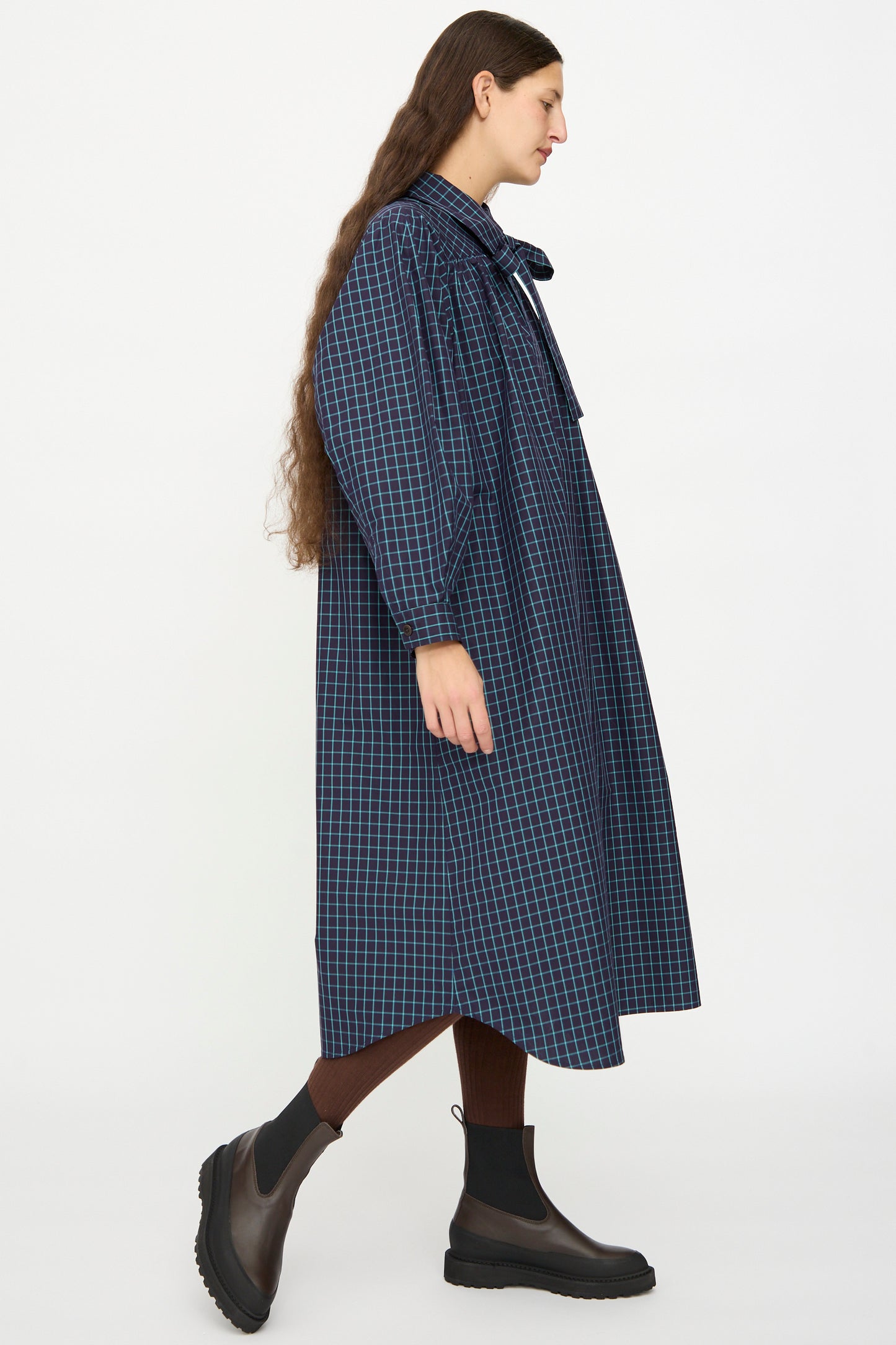 A person with long brown hair wears the KasMaria Cotton Poplin Smock Dress in Blue Check, paired with brown tights and black ankle boots, walking against a plain white background.
