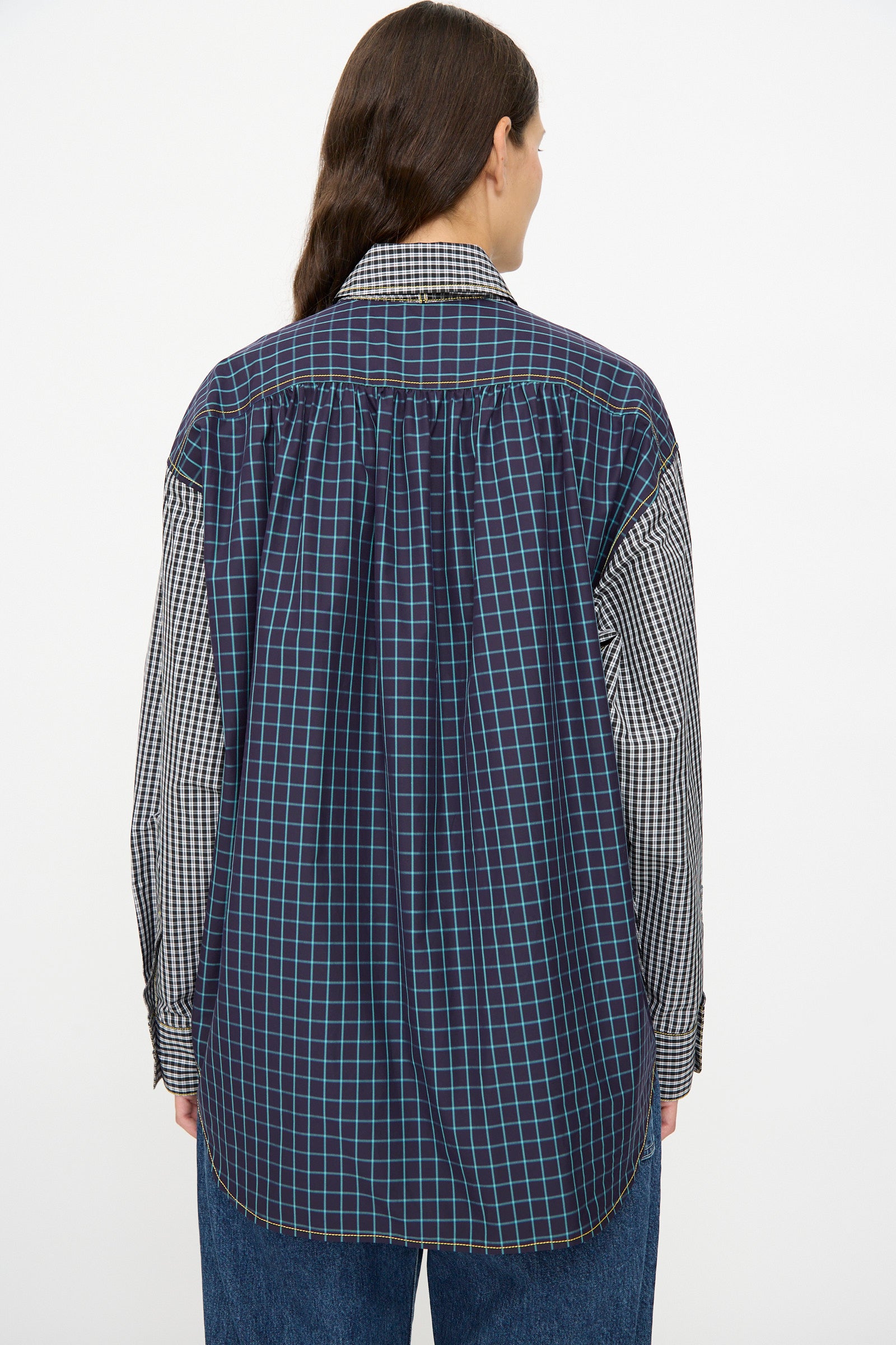 A person stands facing away, wearing the KasMaria Cotton Shirt with Tie in Check Combo—a relaxed fit, long-sleeve button down with navy blue, black, and white plaid patterns—paired with blue jeans.