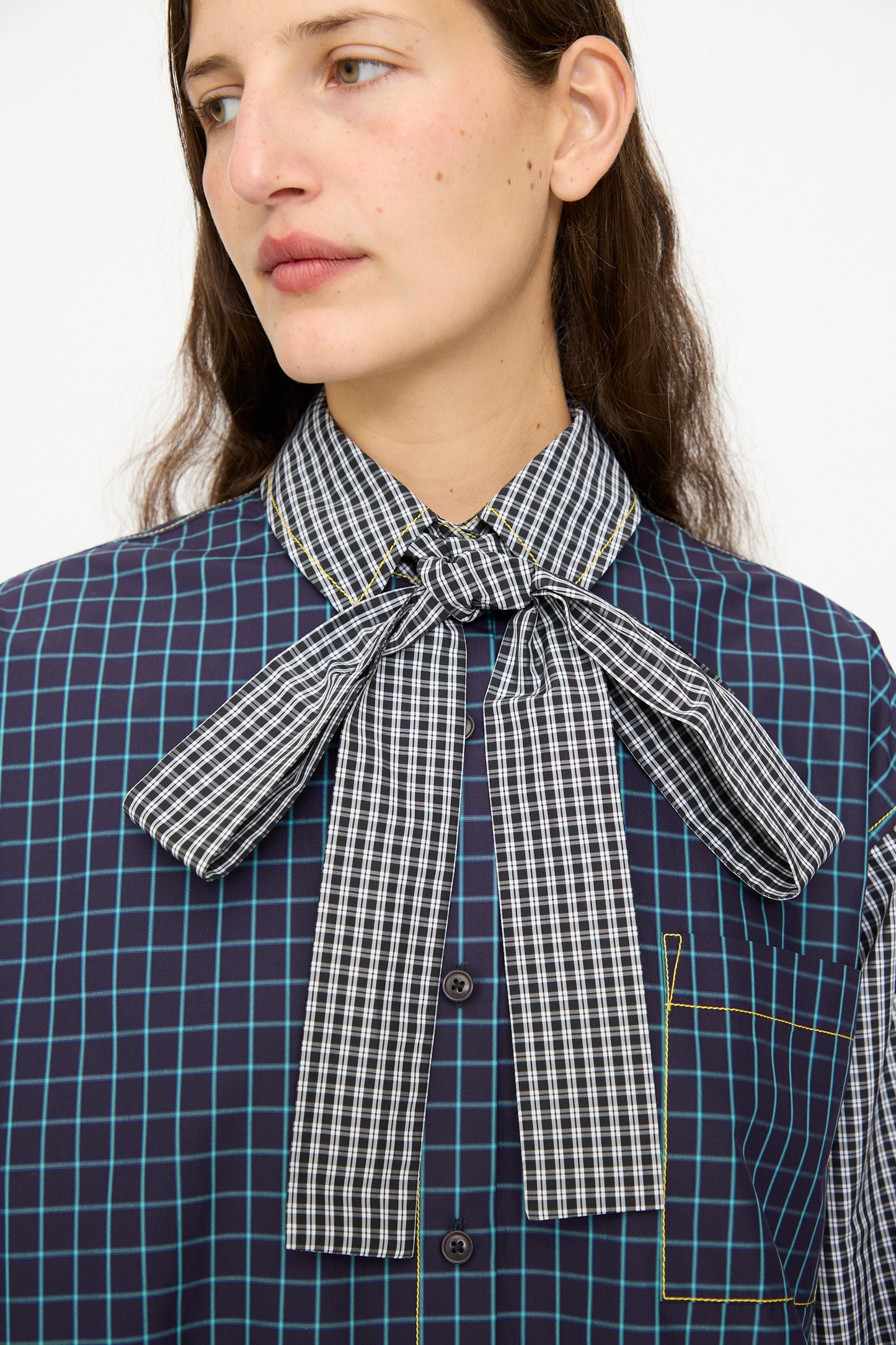 A person wearing the KasMaria Cotton Shirt with Tie in Check Combo, featuring a relaxed fit and blue-green grid pattern shirt with a large black-and-white checkered bow tie at the collar.