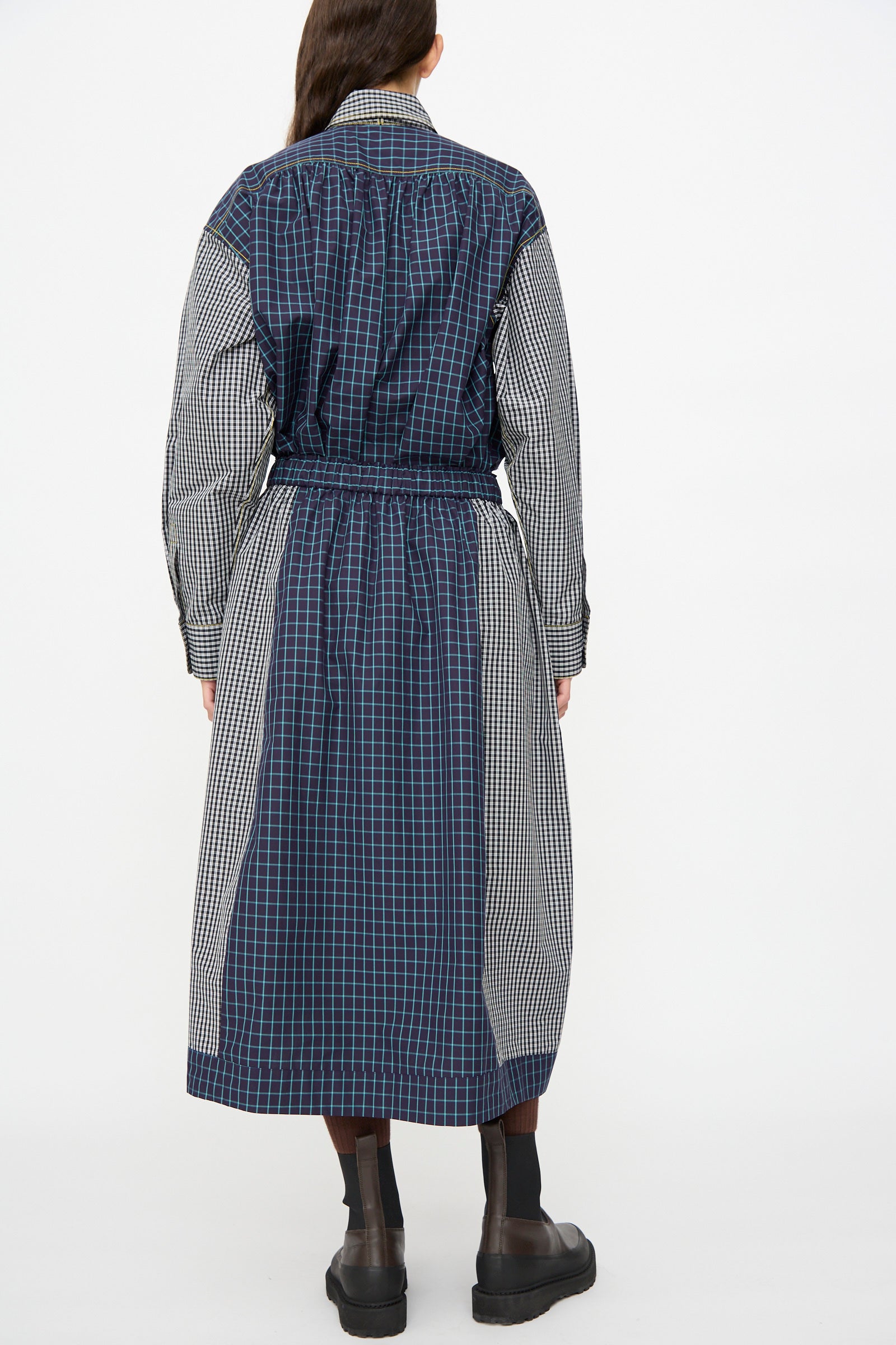 A person stands facing away, wearing the KasMaria Cotton Skirt in Check Combo, featuring mixed blue and black plaid Italian cotton, paired with ankle-high black boots.