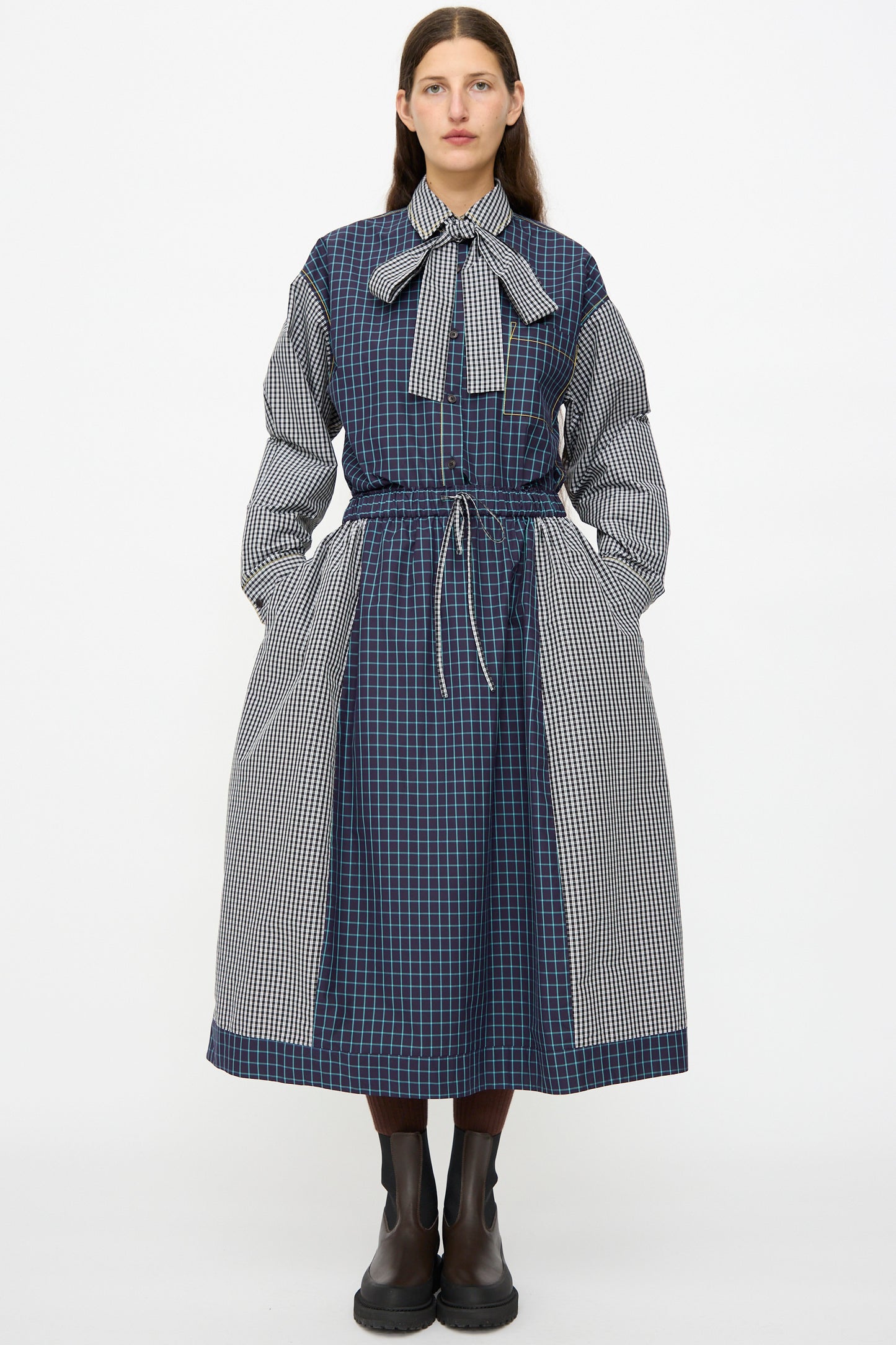 A person faces forward wearing the KasMaria Cotton Skirt in Check Combo—an oversized, long, blue and black checkered Italian cotton skirt—with a bow at the neck and black boots. Hands are in pockets.