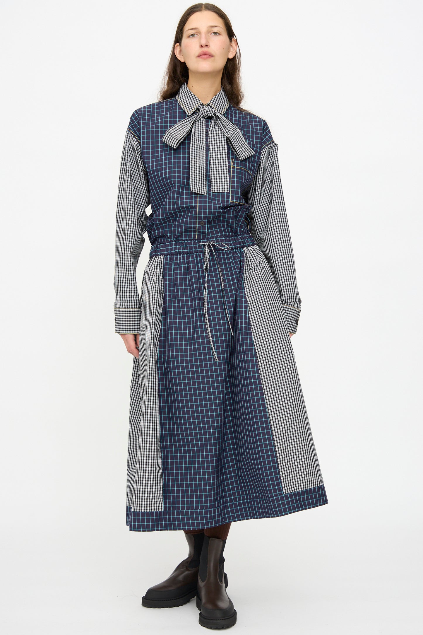 A person stands against a plain background wearing the KasMaria Cotton Skirt in Check Combo, featuring an oversized blue and black check pattern, a bow tie collar, black boots, and made from soft Italian cotton.