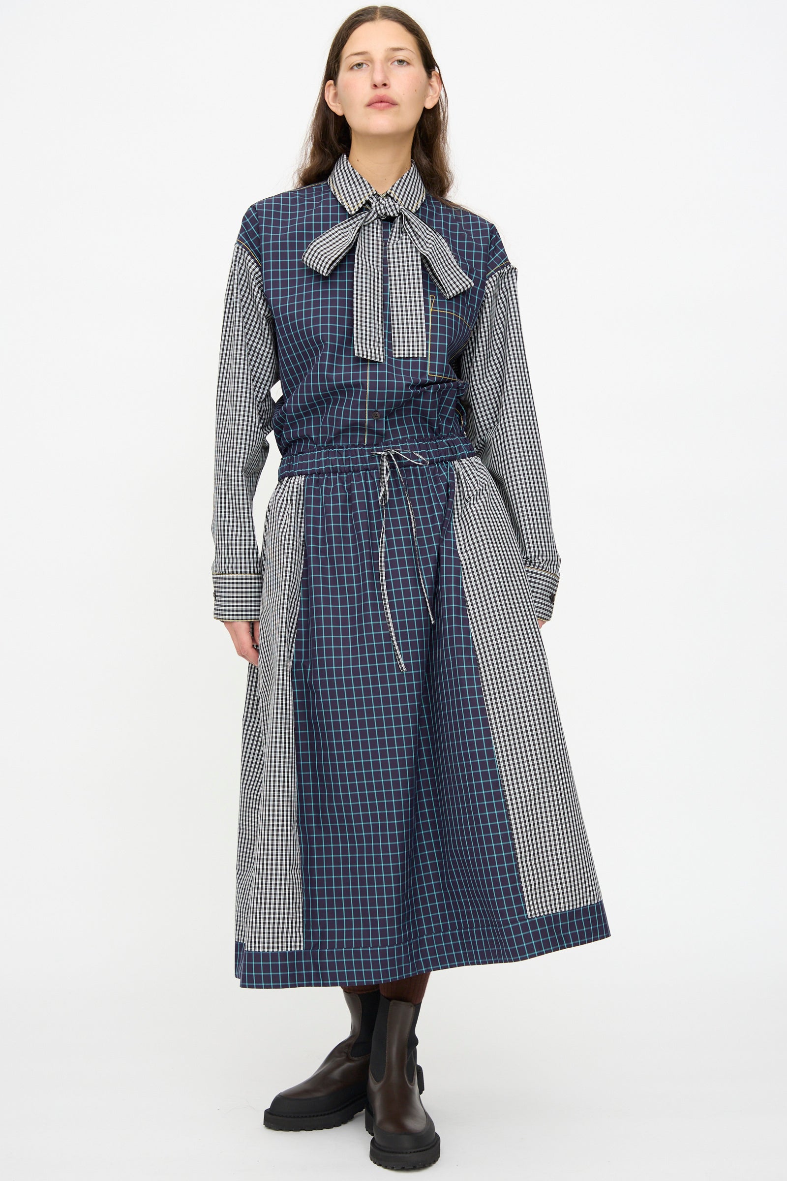 A person stands against a plain background wearing the KasMaria Cotton Skirt in Check Combo, featuring an oversized blue and black check pattern, a bow tie collar, black boots, and made from soft Italian cotton.