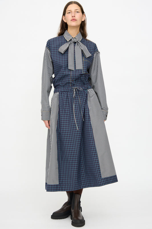 A person stands against a plain background wearing the KasMaria Cotton Skirt in Check Combo, featuring an oversized blue and black check pattern, a bow tie collar, black boots, and made from soft Italian cotton.