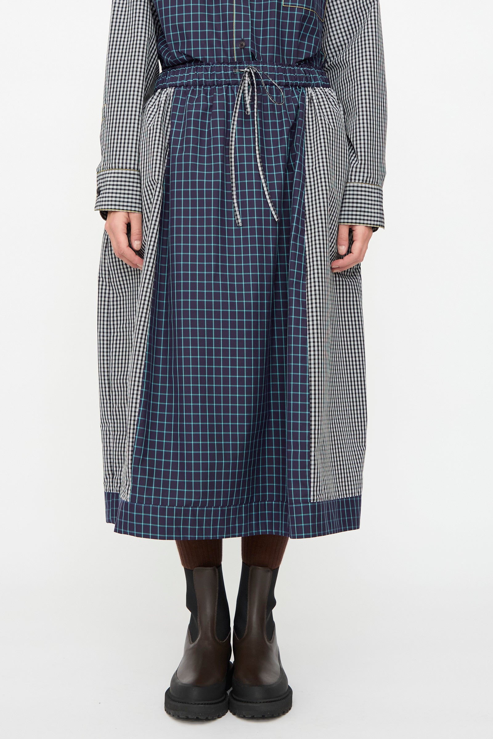 A person wearing the KasMaria Cotton Skirt in Check Combo with brown tights and black boots, standing against a plain white background.