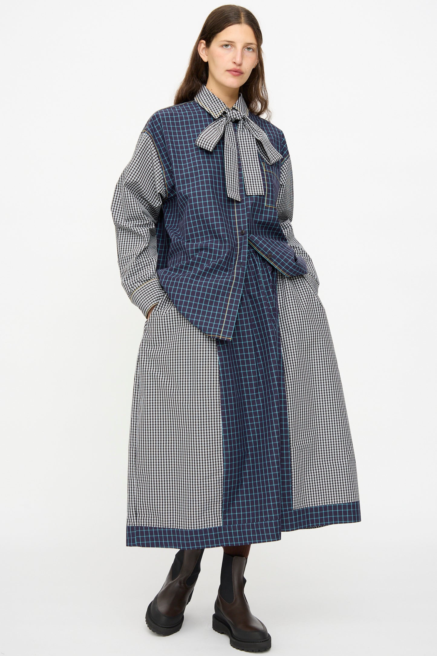 A person wears the KasMaria Cotton Skirt in Check Combo, styled with a long, oversized plaid shirt, a large neck bow, and black ankle boots, standing against a plain white background.