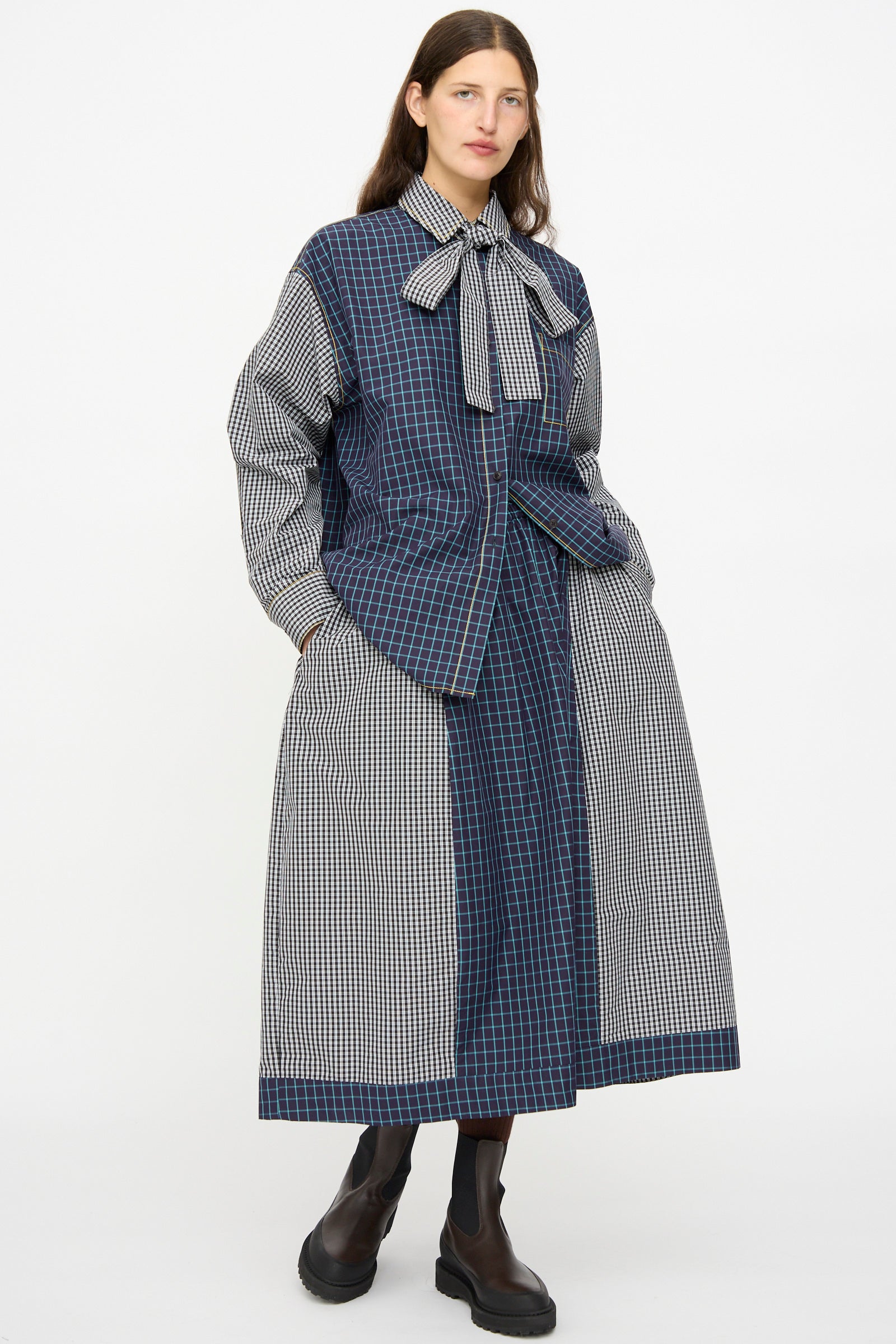 A person wears the KasMaria Cotton Skirt in Check Combo, styled with a long, oversized plaid shirt, a large neck bow, and black ankle boots, standing against a plain white background.