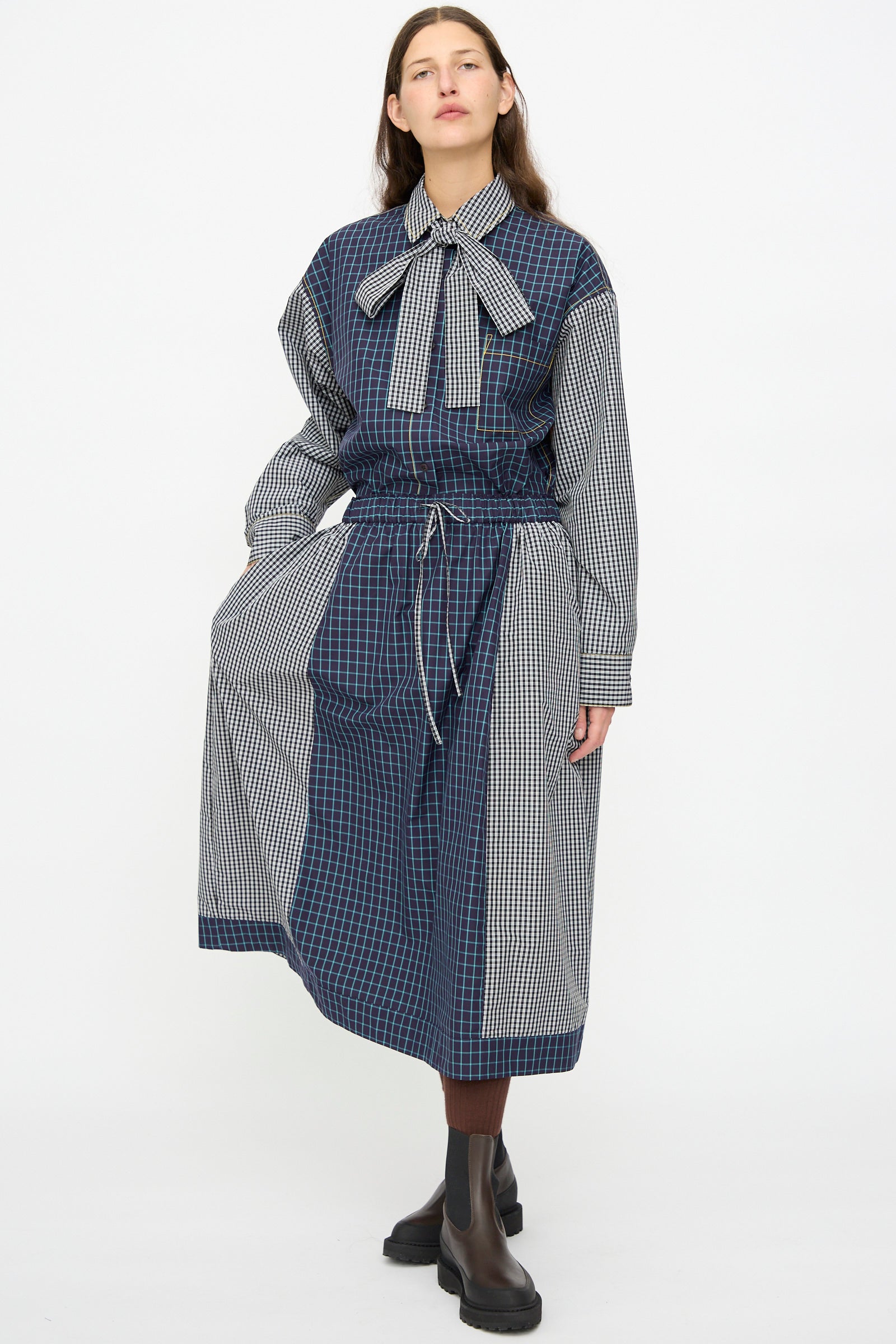 A person wears the KasMaria Cotton Skirt in Check Combo, made from Italian cotton with blue plaid and check patterns, styled with a large bow collar, long sleeves, oversized drawstring waist, brown socks, and black boots.