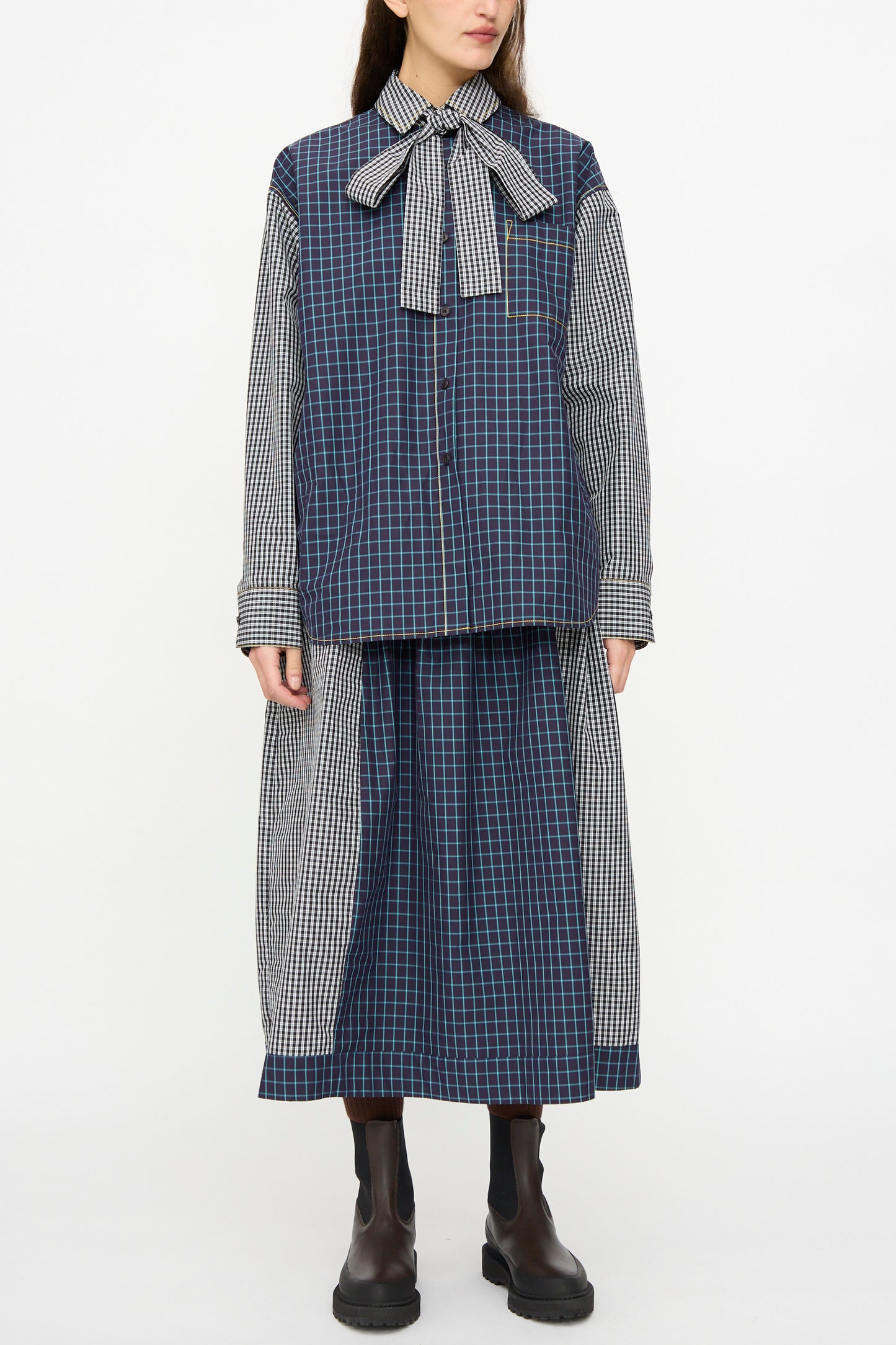 A person wears the KasMaria Cotton Skirt in Check Combo, featuring an oversized fit, large neck bow, long sleeves, matching midi skirt, and black ankle boots, standing against a plain white background.