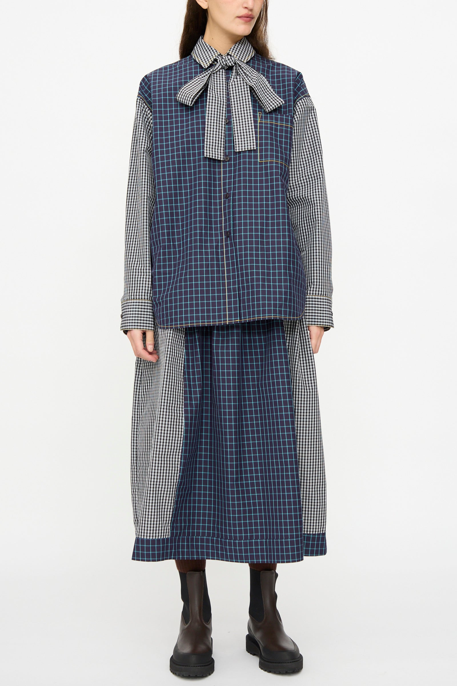 A person wears the KasMaria Cotton Skirt in Check Combo, featuring an oversized fit, large neck bow, long sleeves, matching midi skirt, and black ankle boots, standing against a plain white background.