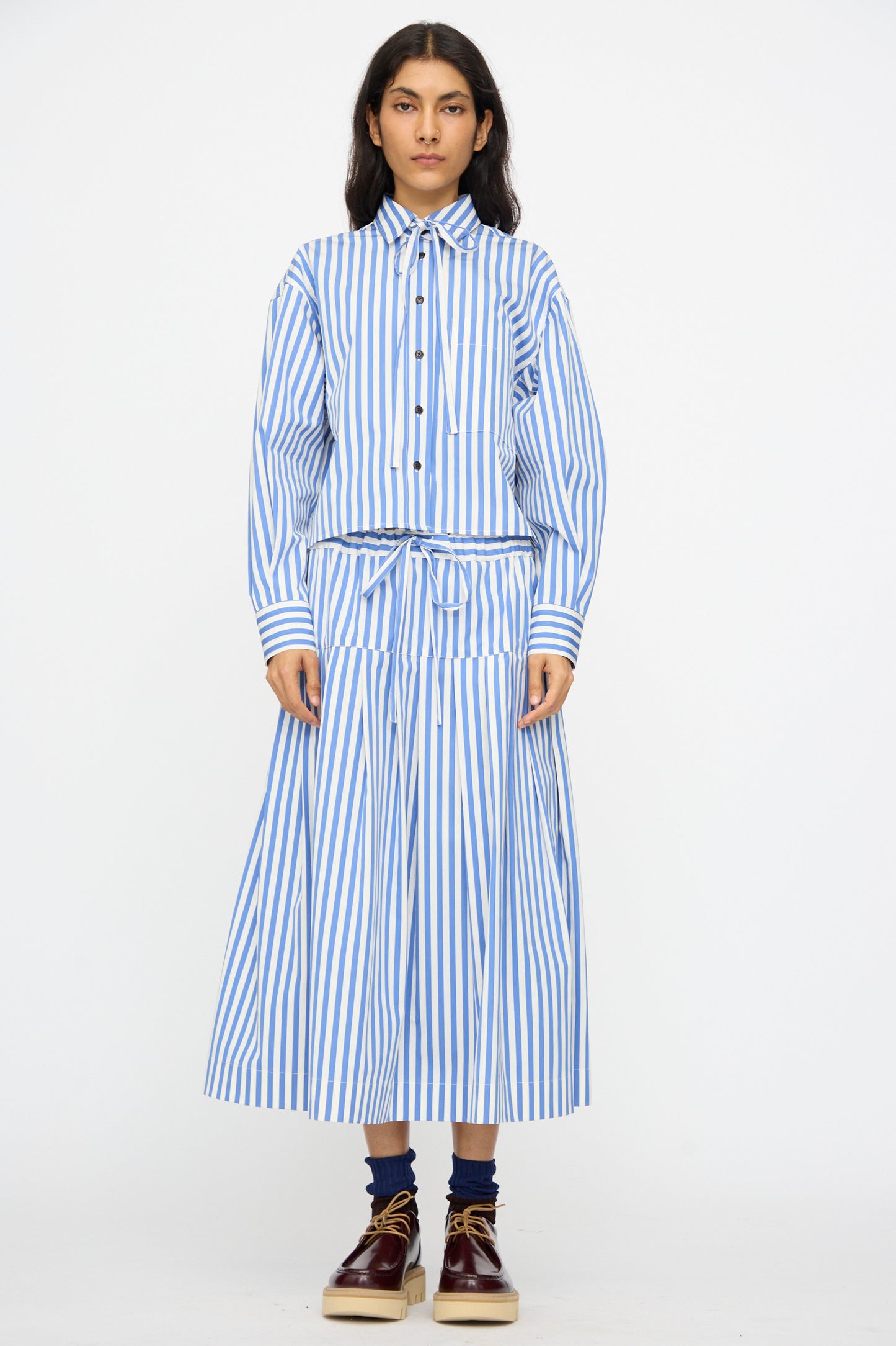 A person faces forward wearing the KasMaria Cotton Stripe Pleated Skirt in Blue, styled with a matching blue and white striped shirt, dark socks, and beige platform shoes.