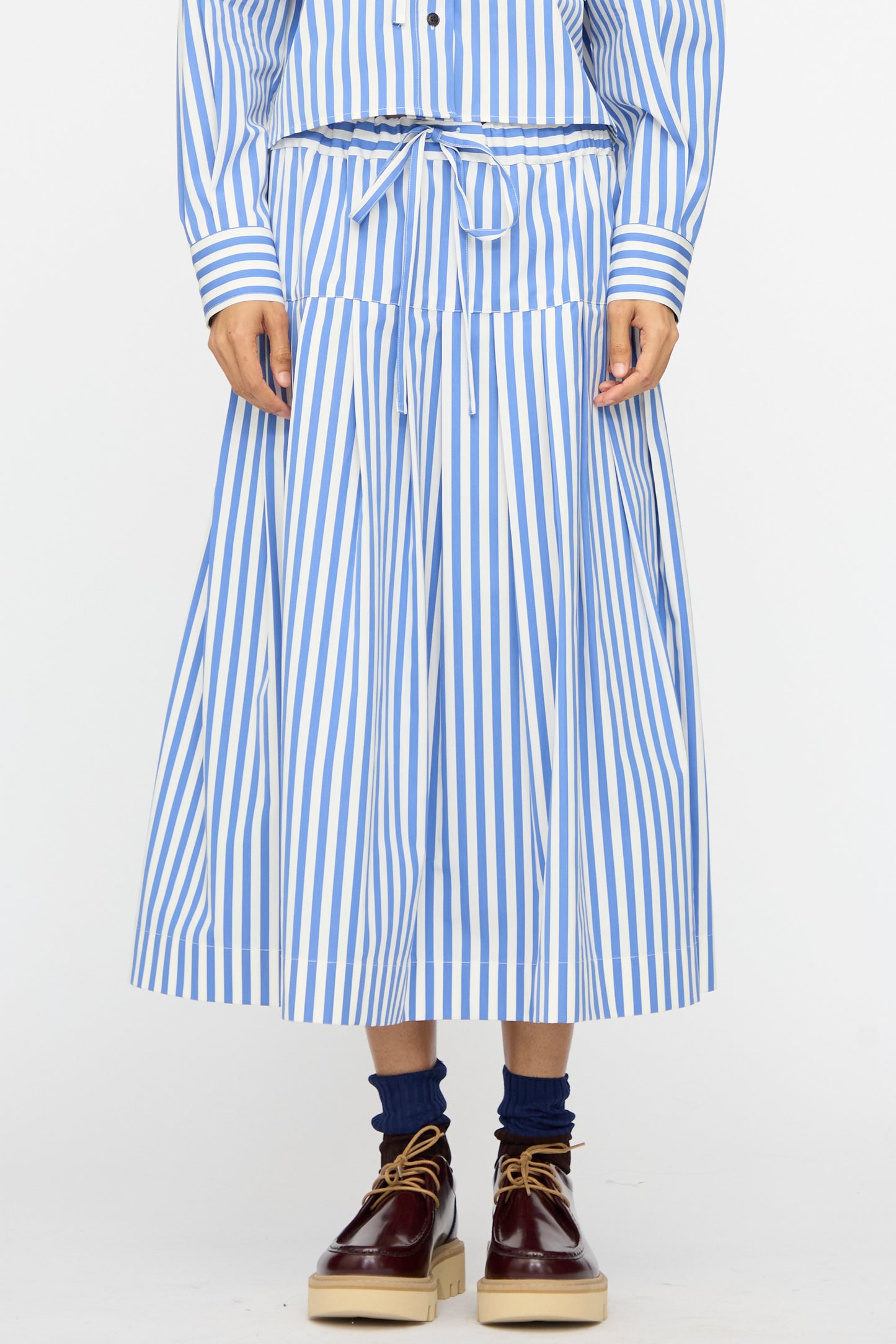 Person wearing the Cotton Stripe Pleated Skirt in Blue from KasMaria, styled with a matching drawstring top, navy socks, and brown platform shoes.