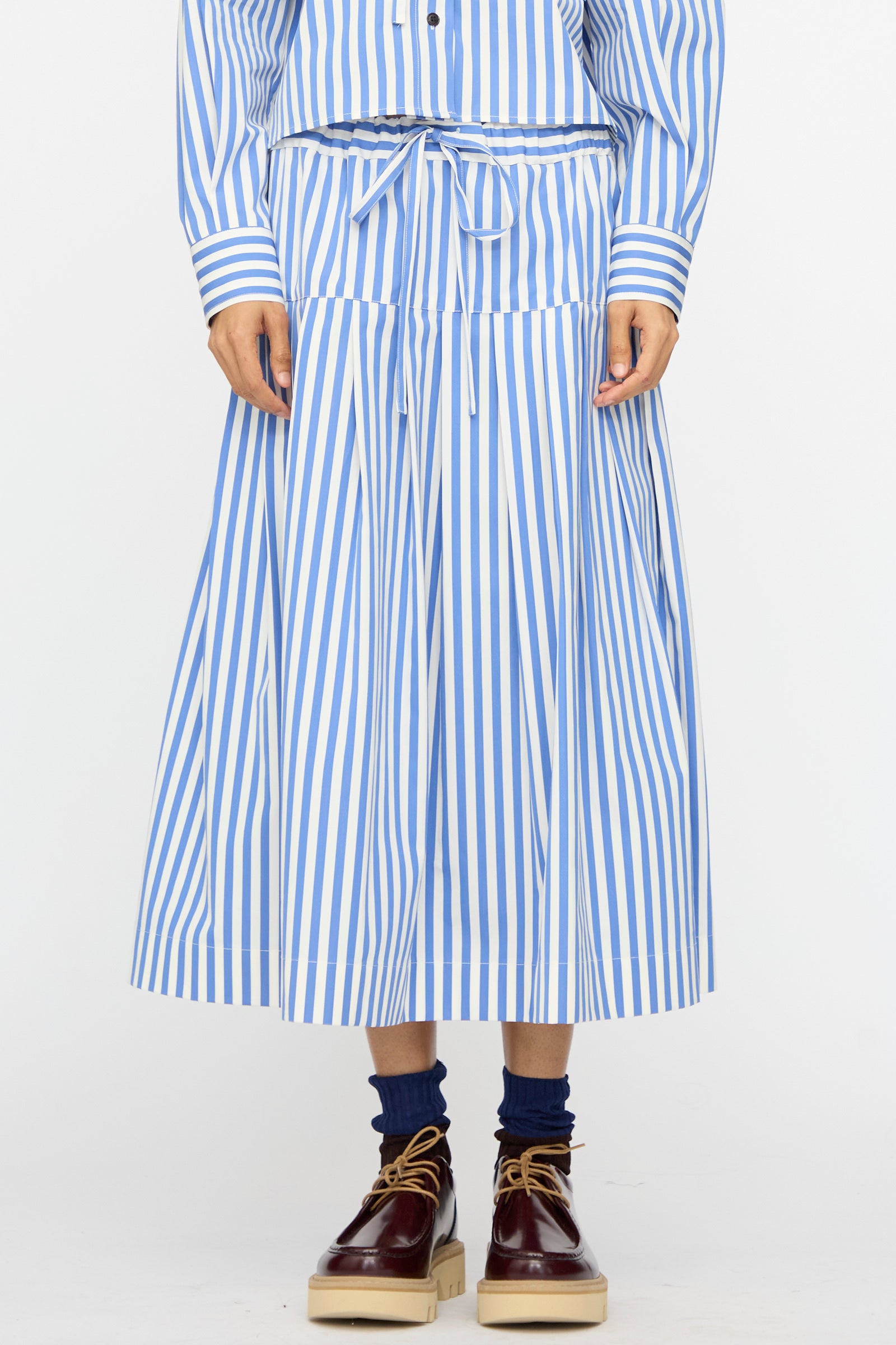 Person wearing the Cotton Stripe Pleated Skirt in Blue from KasMaria, styled with a matching drawstring top, navy socks, and brown platform shoes.