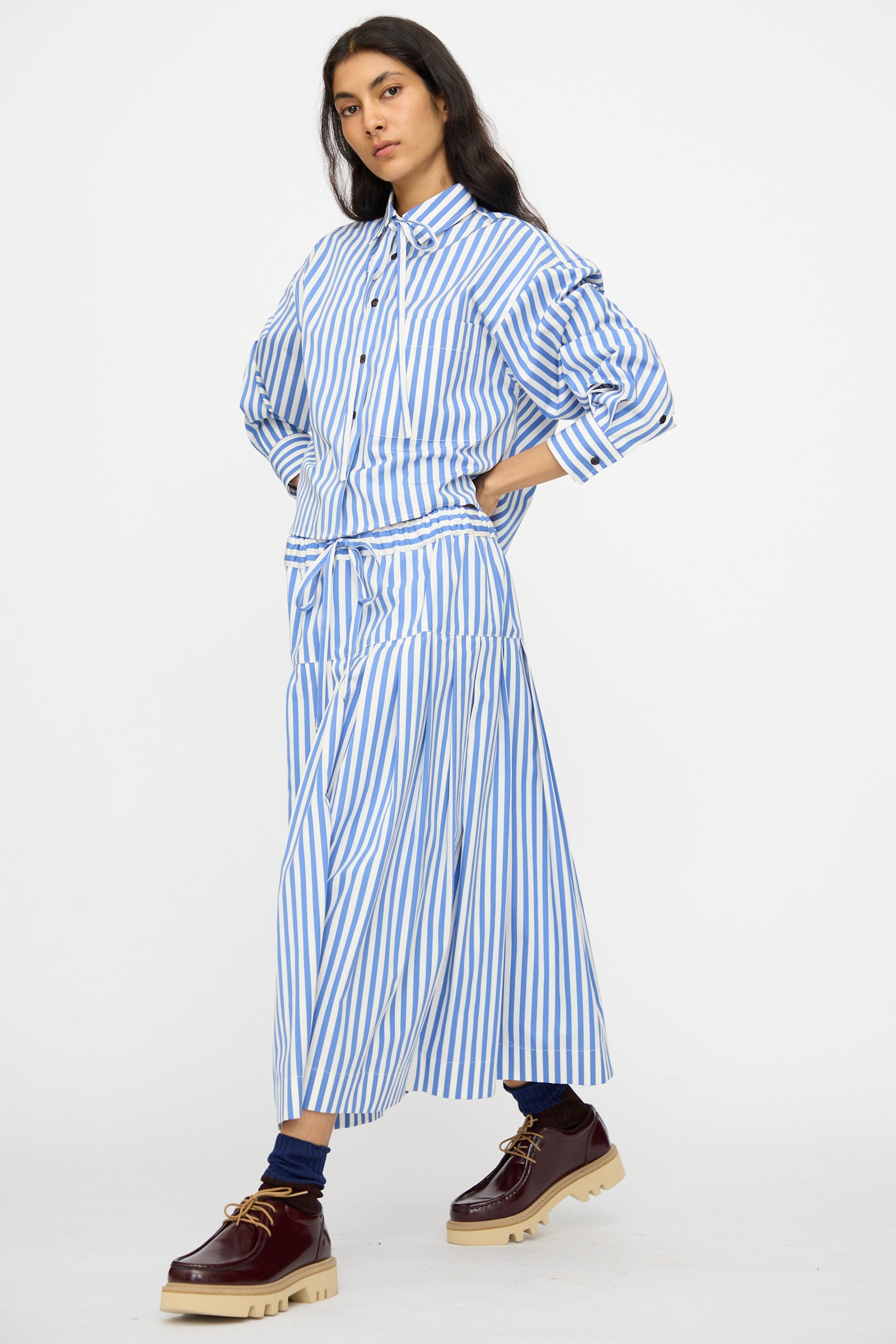 Model wearing the KasMaria Cotton Stripe Pleated Skirt in Blue with a matching shirt and brown chunky shoes, standing against a plain white background.