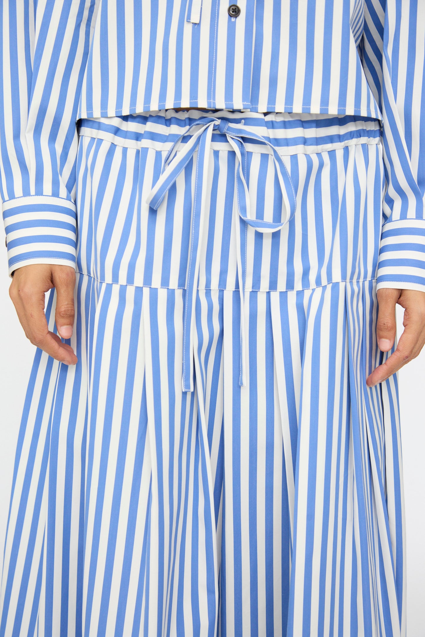 A person is seen wearing the KasMaria Cotton Stripe Pleated Skirt in Blue, featuring vertical stripes, an A-line silhouette, a drawstring waist, and pleats; only their torso and hands are visible.