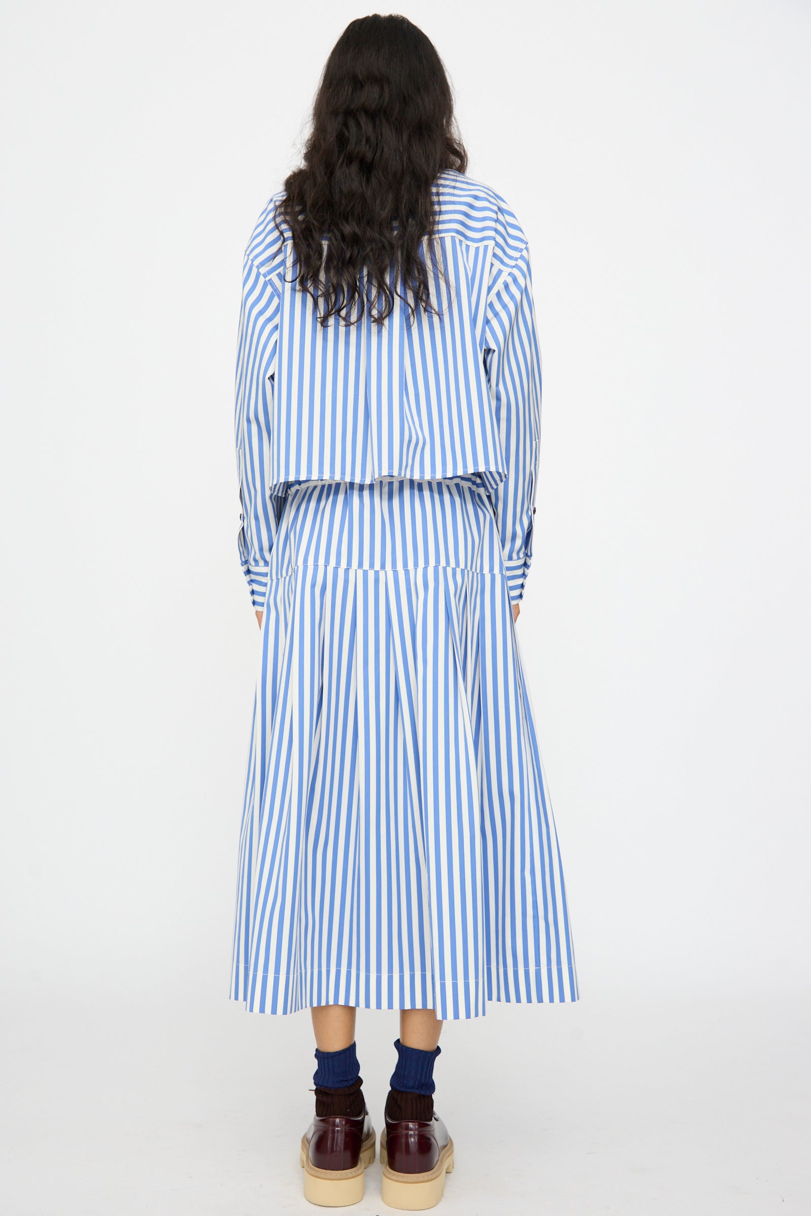 Person standing with back to camera, wearing the KasMaria Cotton Stripe Pleated Skirt in Blue, paired with dark socks and brown shoes.
