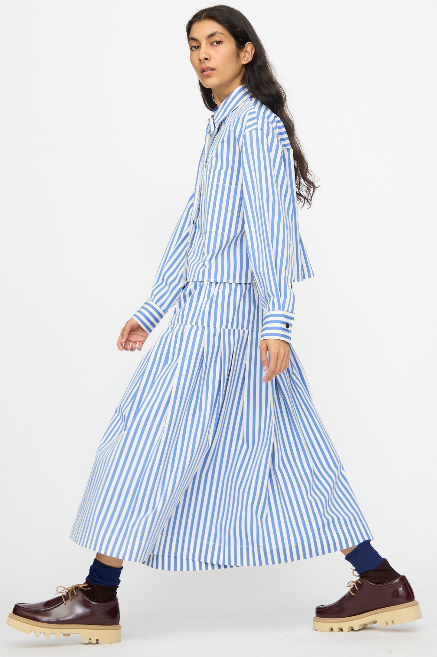 A person in a KasMaria Cotton Stripe Pleated Skirt in Blue with a matching top, dark socks, and brown platform shoes walks against a plain background.