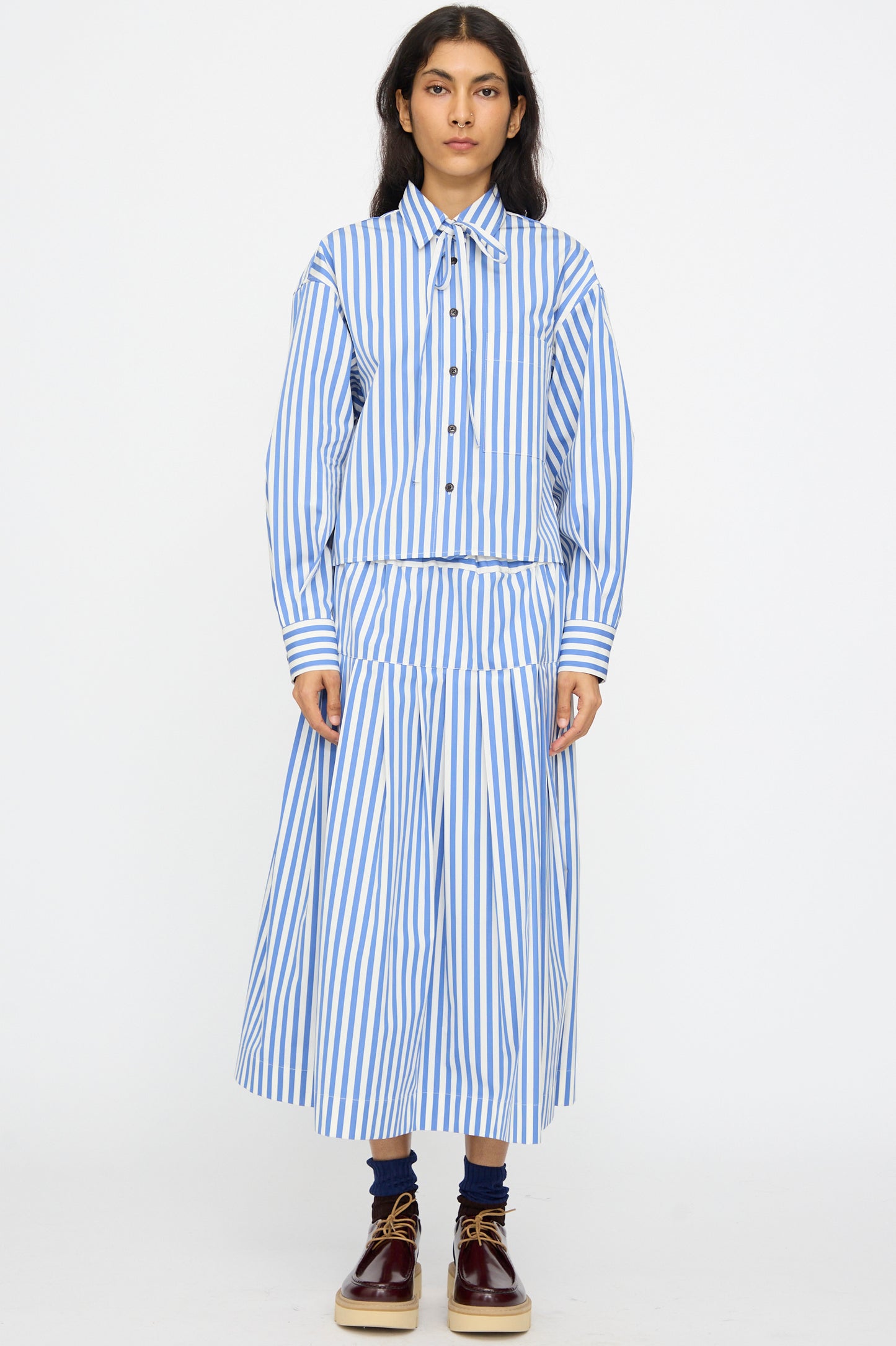 A person wears the KasMaria Cotton Stripe Shirt with Tie in Blue, featuring a cropped boxy fit, paired with a matching skirt, burgundy shoes, and dark socks against a plain white background.