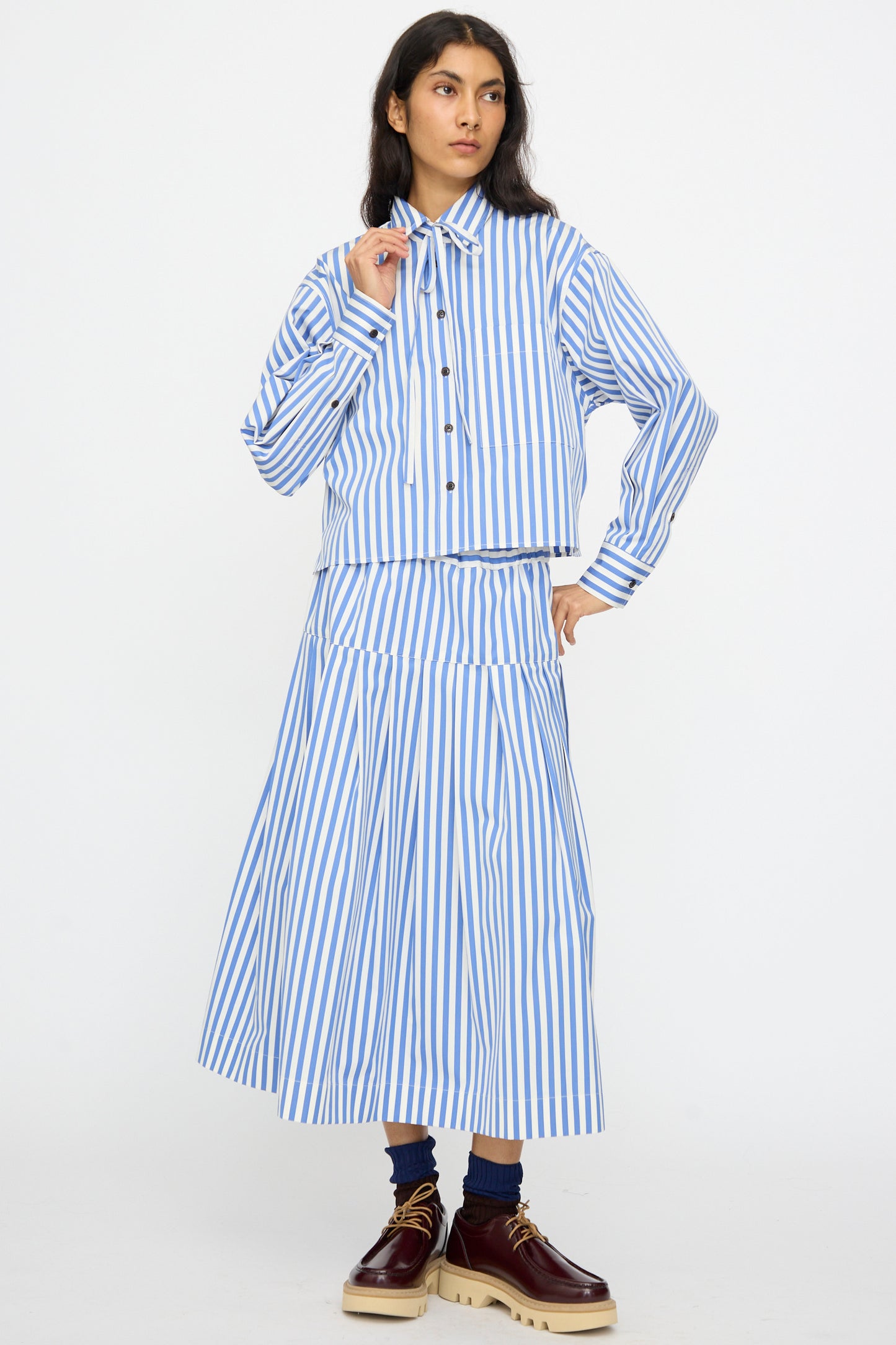 Person standing against a plain background wearing the KasMaria Cotton Stripe Shirt with Tie in Blue, featuring a cropped boxy fit, matching midi skirt, maroon shoes, and blue socks.