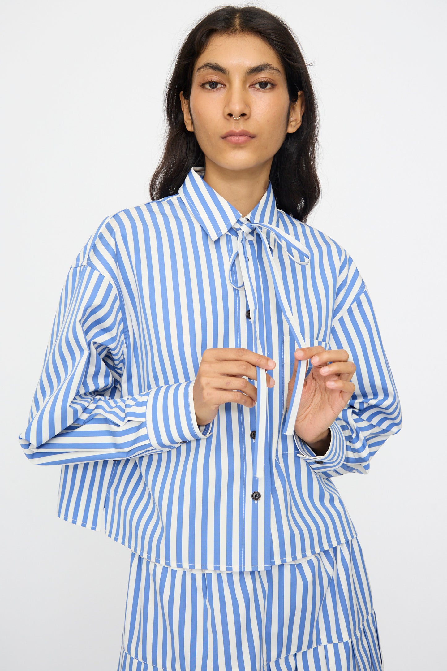 A person models the KasMaria Cotton Stripe Shirt with Tie in Blue, featuring a cropped boxy fit and blue-and-white stripes, against a plain background.