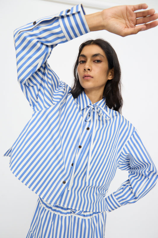 A person wears the KasMaria Cotton Stripe Shirt with Tie in Blue, featuring blue and white vertical stripes and a cropped boxy fit, standing with one arm raised and the other on their hip against a plain background. Made in NYC.