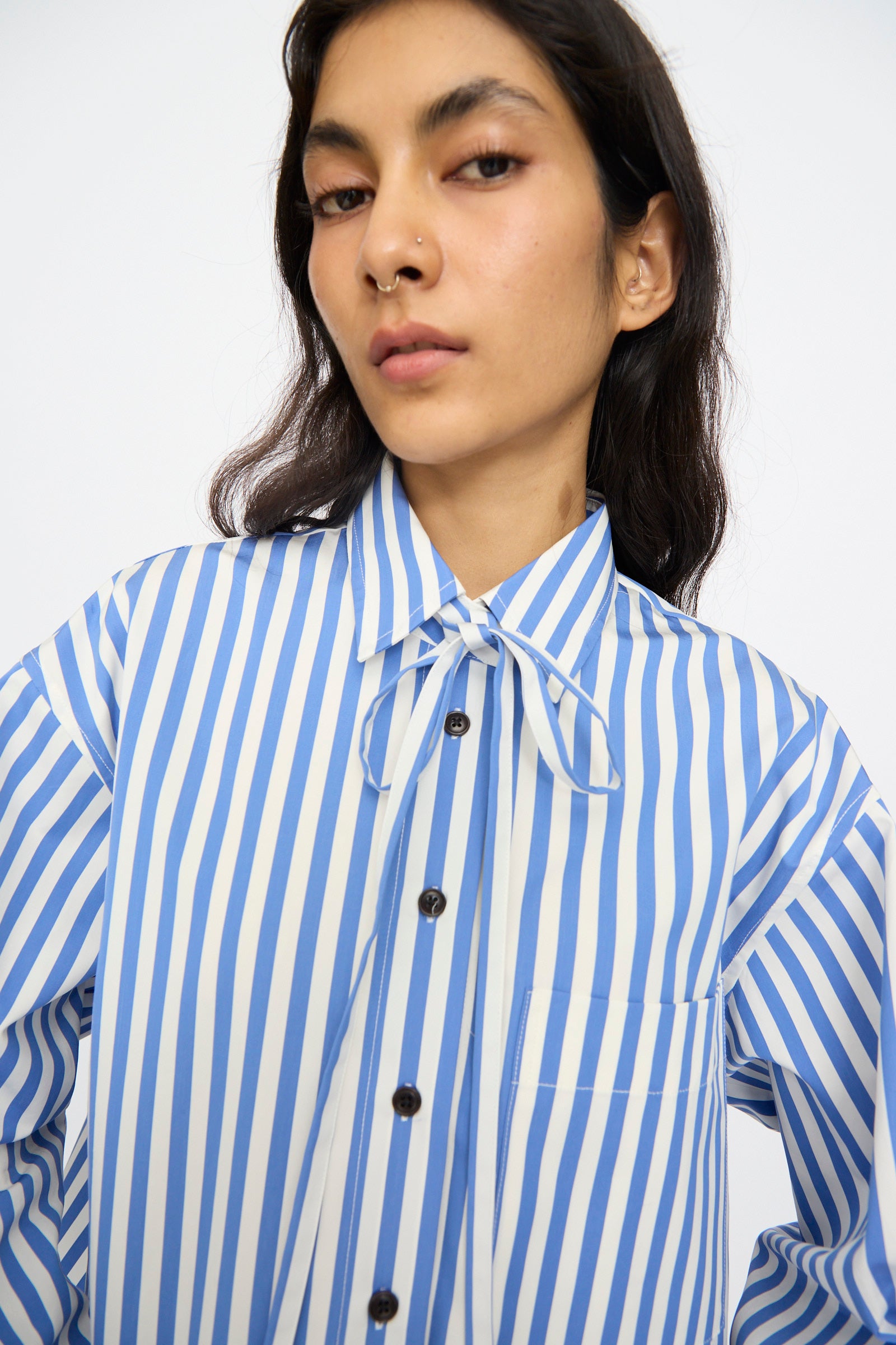 A person wears the KasMaria Cotton Stripe Shirt with Tie in Blue, featuring blue and white vertical stripes, a front pocket, bow collar, cropped boxy fit, and posing against a plain background. Made in NYC.