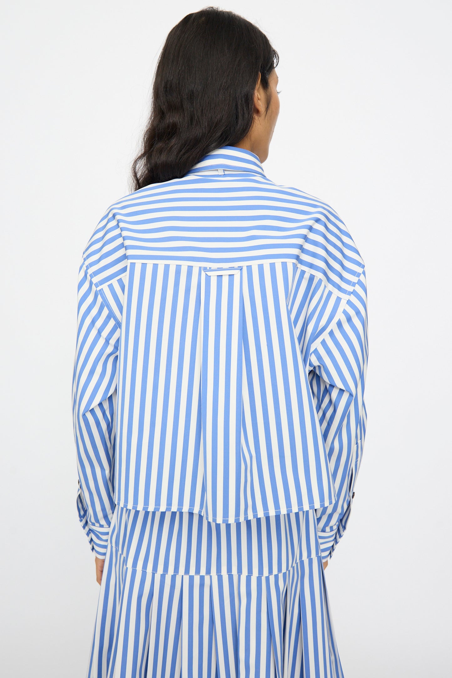 A person with long dark hair is seen from behind wearing the KasMaria Cotton Stripe Shirt with Tie in Blue, featuring a boxy cropped fit and blue and white vertical stripes, against a plain background.