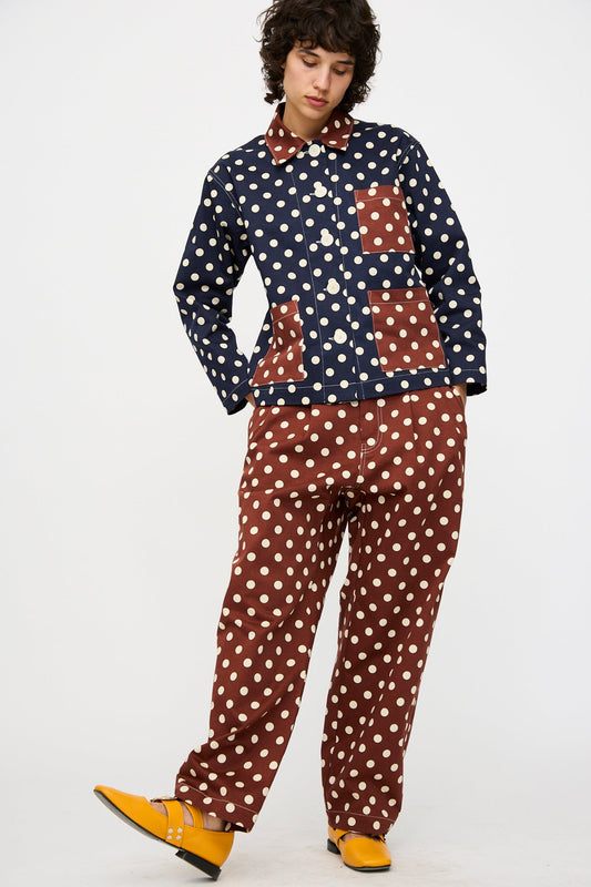 A person stands against a plain white background wearing KasMaria's Cotton Twill Pleated Trouser in Polka Dot, paired with a navy and brown polka-dot matching set and yellow shoes.