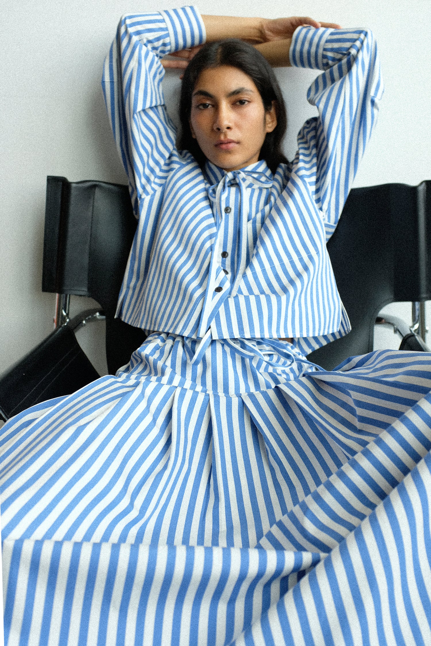 A woman sitting on a black chair with her arms up wearing KasMaria's Cotton Stripe Shirt with Tie in Blue, and the Cotton Stripe Pleated Skirt in Blue.