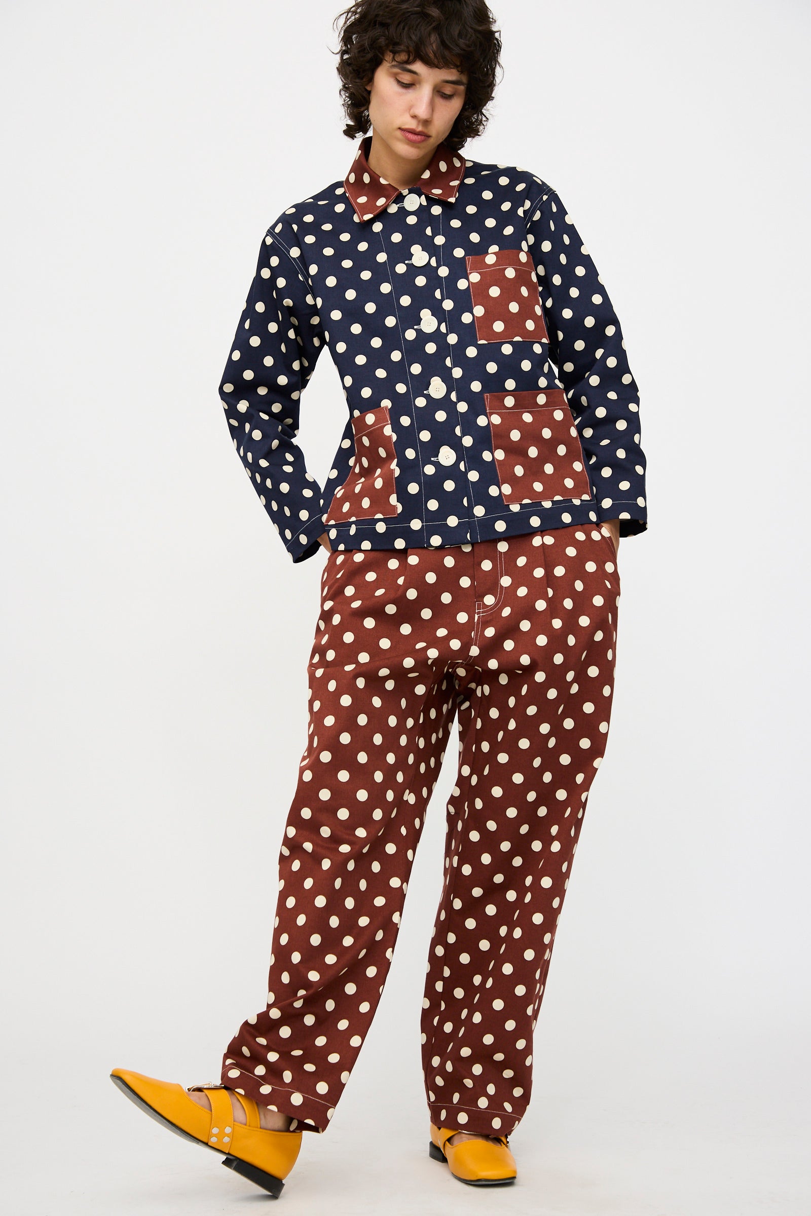 A person wearing KasMaria's Cotton Twill Pleated Trouser in Polka Dot with a navy and brown jacket and yellow shoes stands against a plain white background.