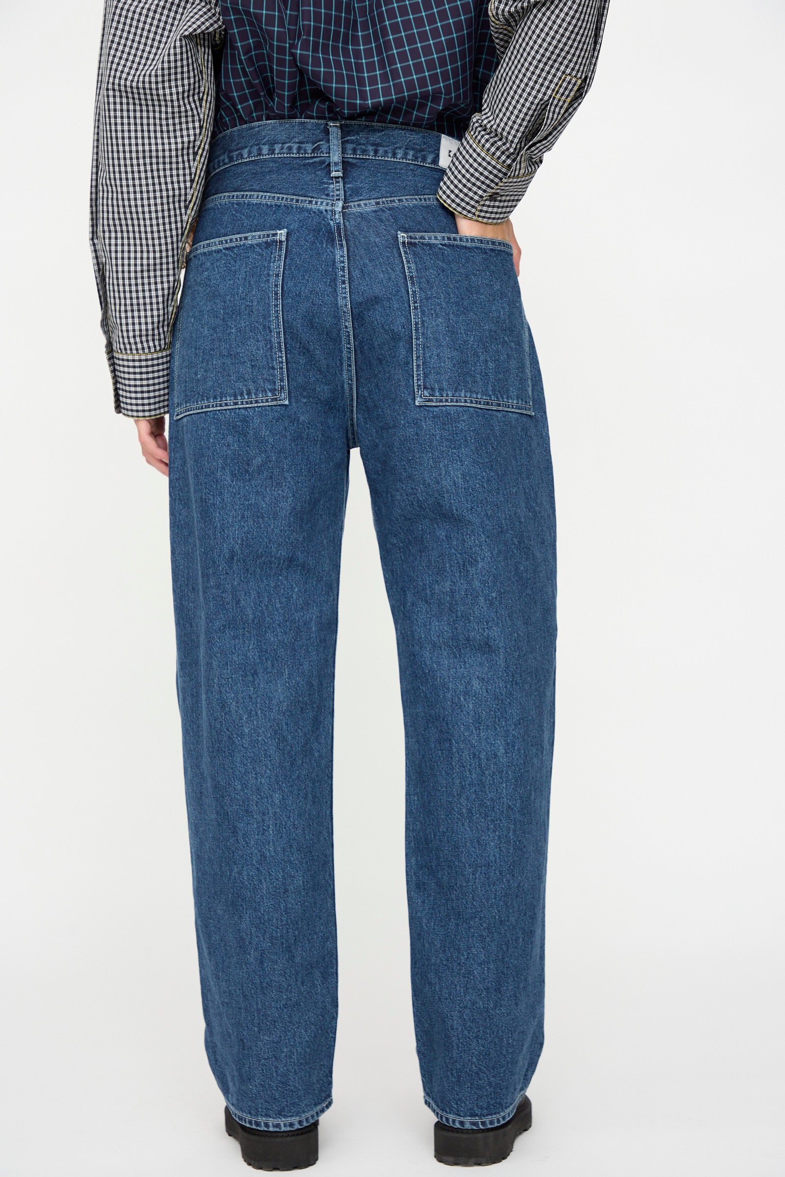 A person wearing KasMaria’s Japanese Denim Pleated Jean in Indigo, featuring a high-waisted, loose fit and two large back pockets, is photographed from behind against a plain background.