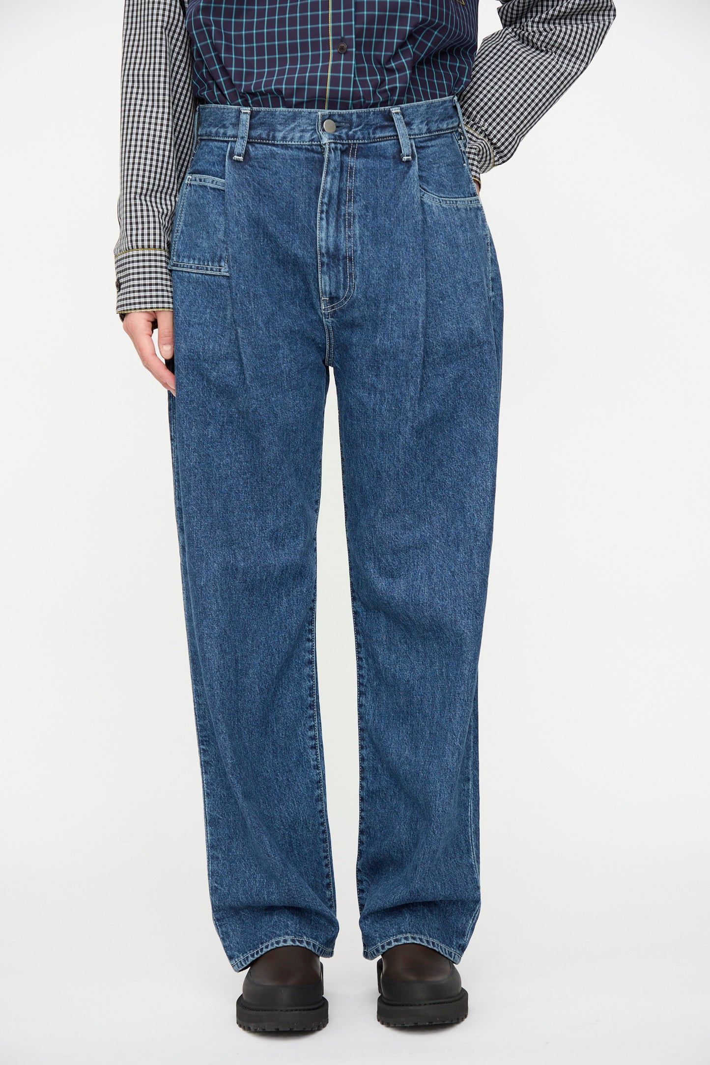 A person wears KasMaria's Japanese Denim Pleated Jean in Indigo with a multicolored checkered shirt and black shoes, standing against a plain background.