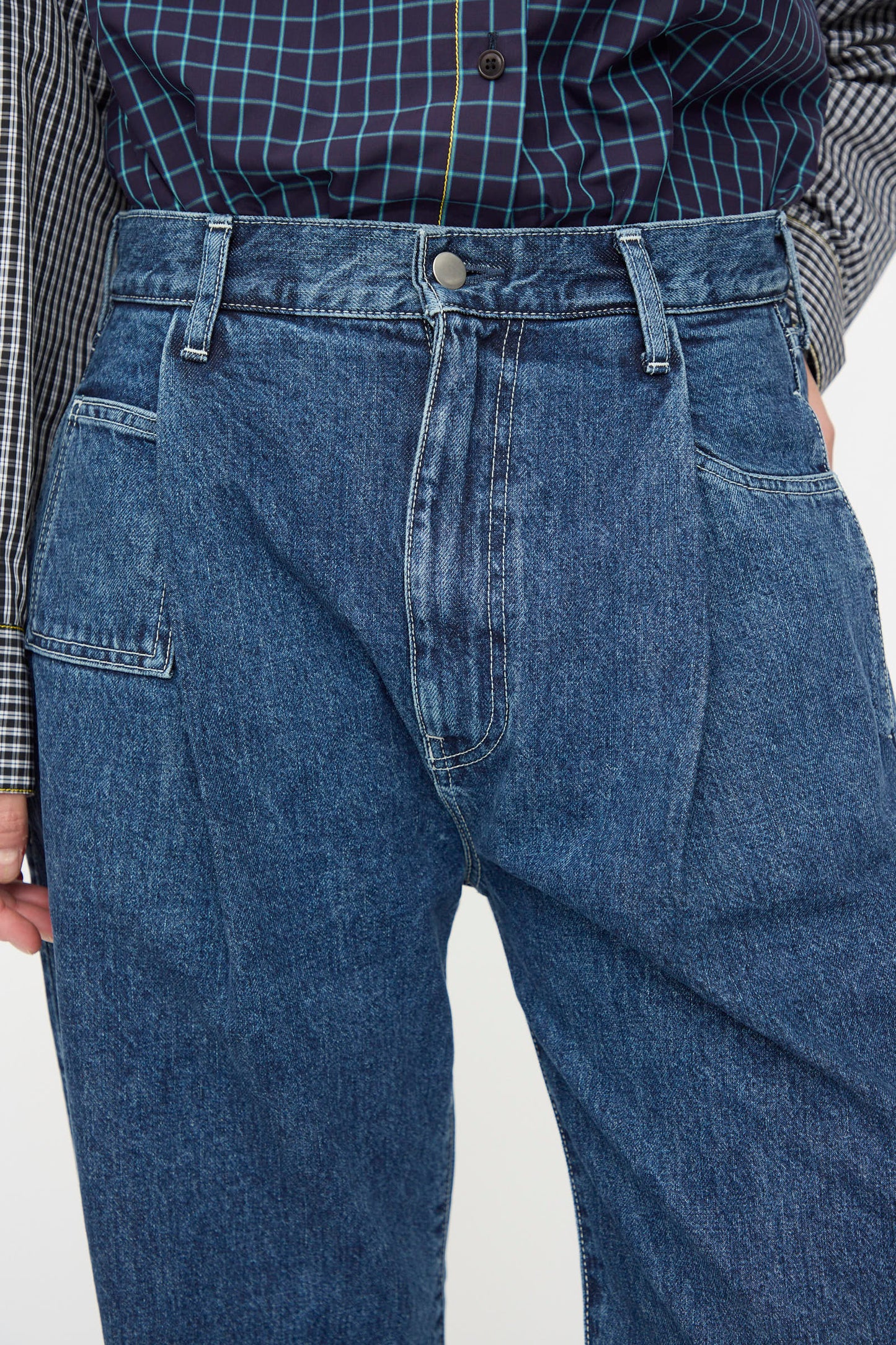 A person wearing KasMaria's Japanese Denim Pleated Jean in Indigo with a button fly and a plaid shirt tucked in.