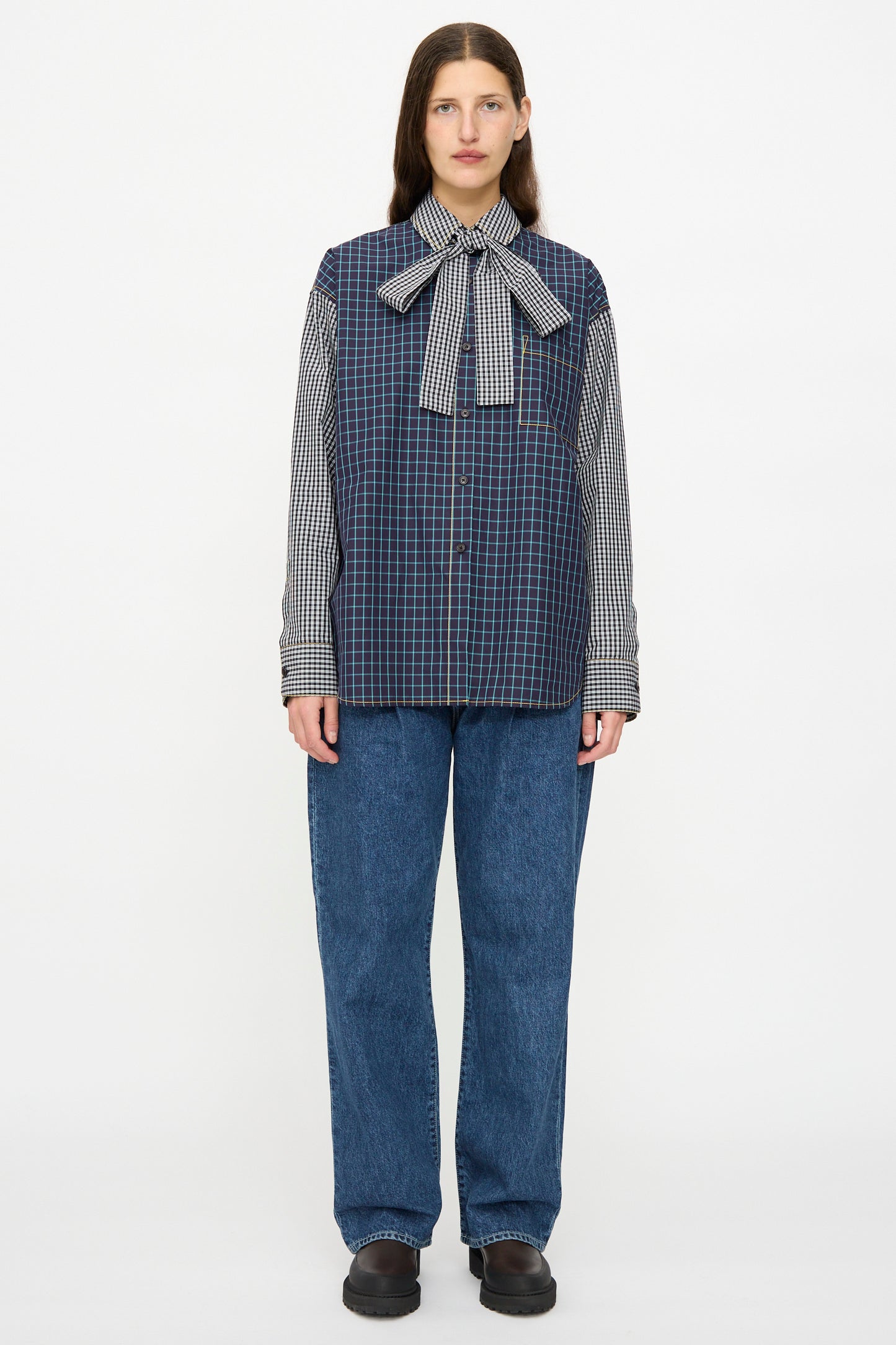 Person stands against a plain background, wearing a plaid shirt with a checkered bow tie, KasMaria Japanese Denim Pleated Jean in Indigo, and black shoes.