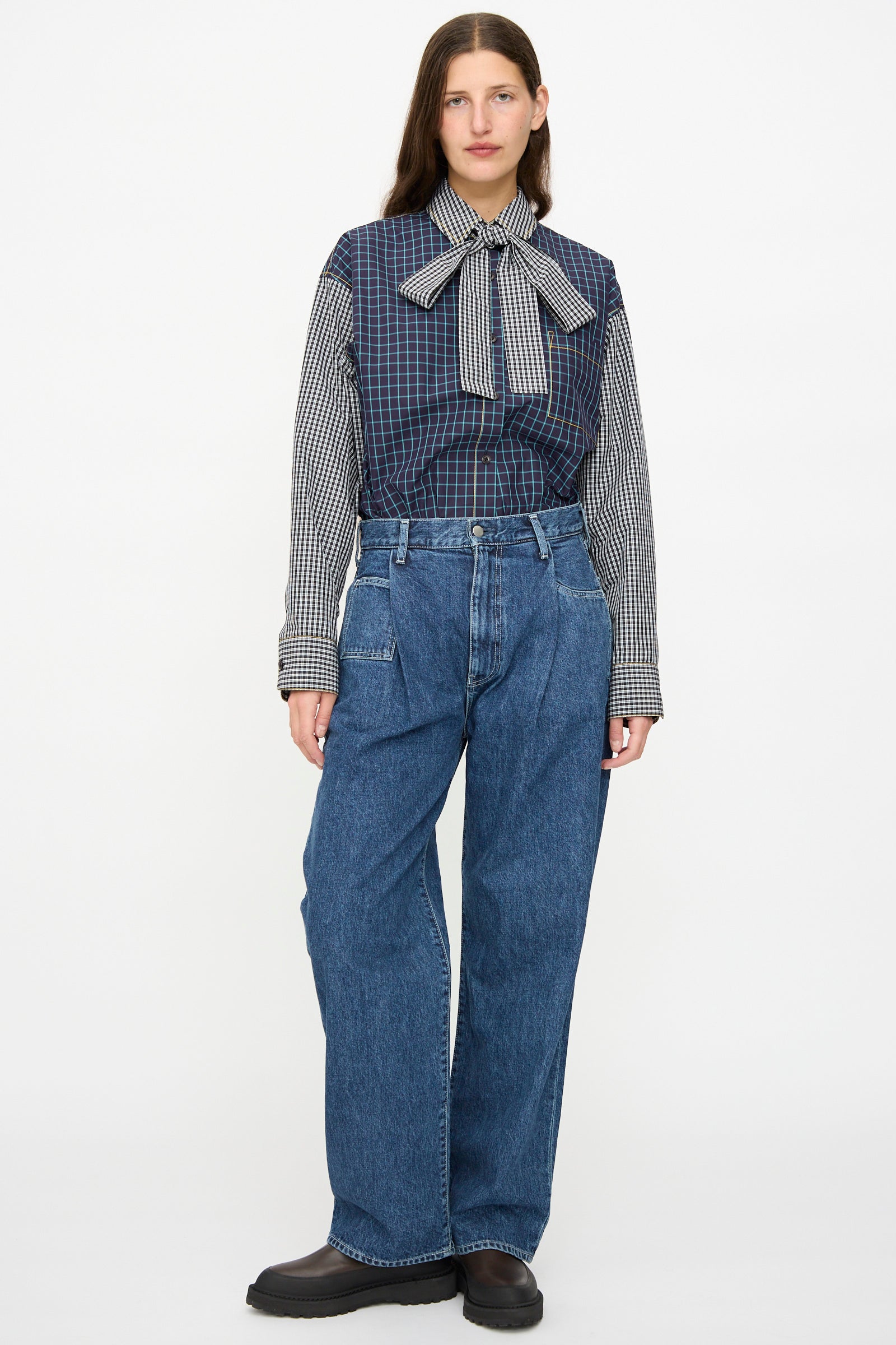 A woman faces forward wearing a long-sleeve checkered blouse with a large bow and KasMaria Japanese Denim Pleated Jean in Indigo, paired with black shoes against a plain background.