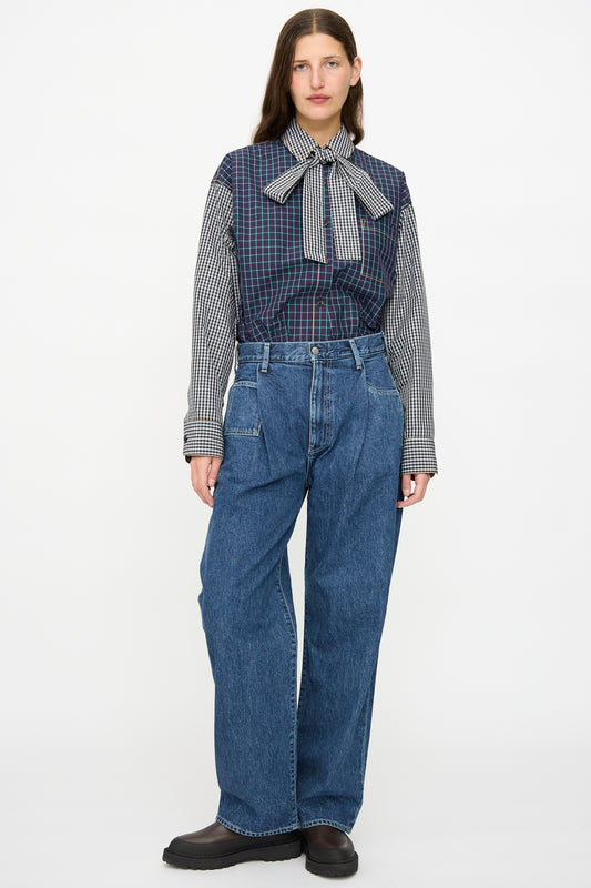 A woman faces forward wearing a long-sleeve checkered blouse with a large bow and KasMaria Japanese Denim Pleated Jean in Indigo, paired with black shoes against a plain background.