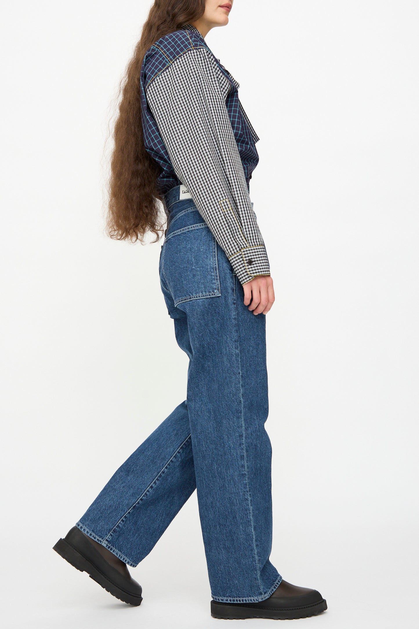 A person wearing a long-sleeve checkered shirt, KasMaria Japanese Denim Pleated Jean in Indigo, and black shoes stands sideways against a plain white background.