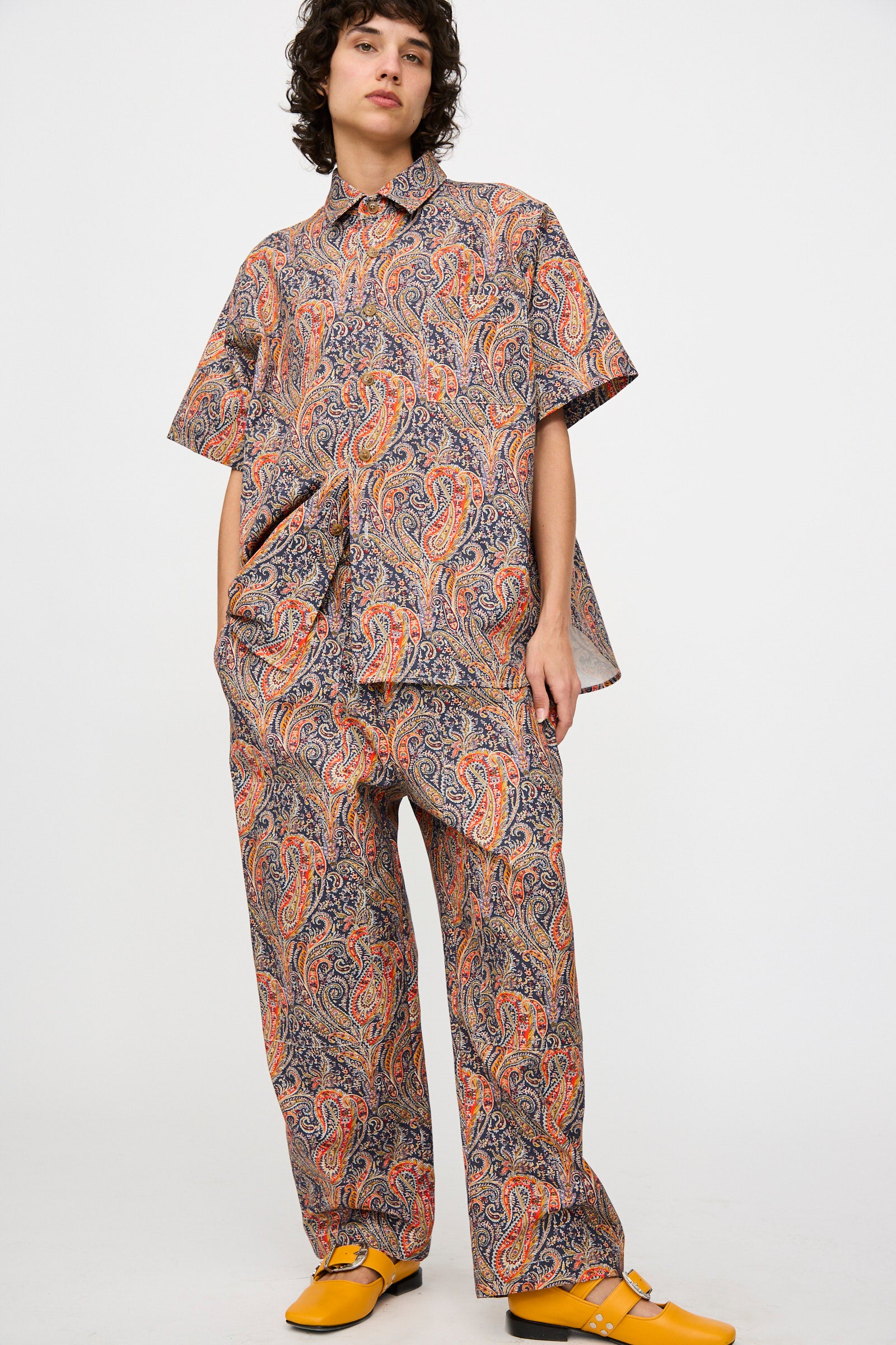 A person stands against a plain background wearing the KasMaria Liberty Cotton Canvas Work Pant in Paisley with a matching paisley short-sleeve shirt, completed with yellow shoes.