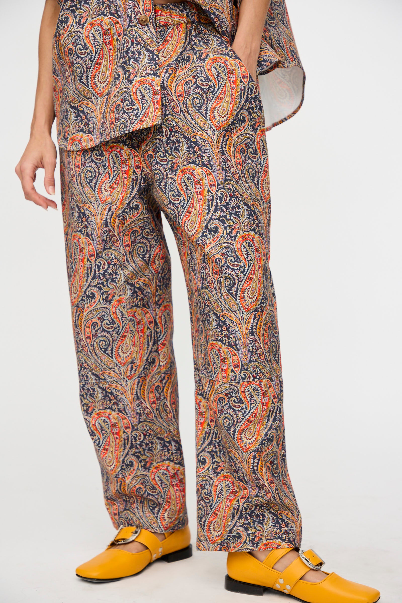 A person wearing the KasMaria Liberty Cotton Canvas Work Pant in Paisley, paired with a matching top and bright yellow Mary Jane shoes, stands against a plain white background.