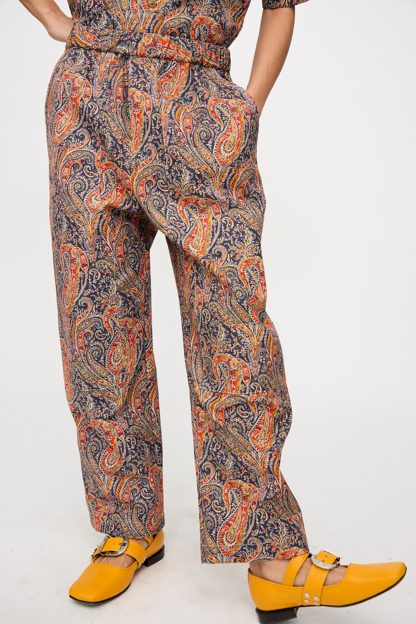 A person models the KasMaria Liberty Cotton Canvas Work Pant in Paisley with yellow buckled shoes, standing against a plain white background.