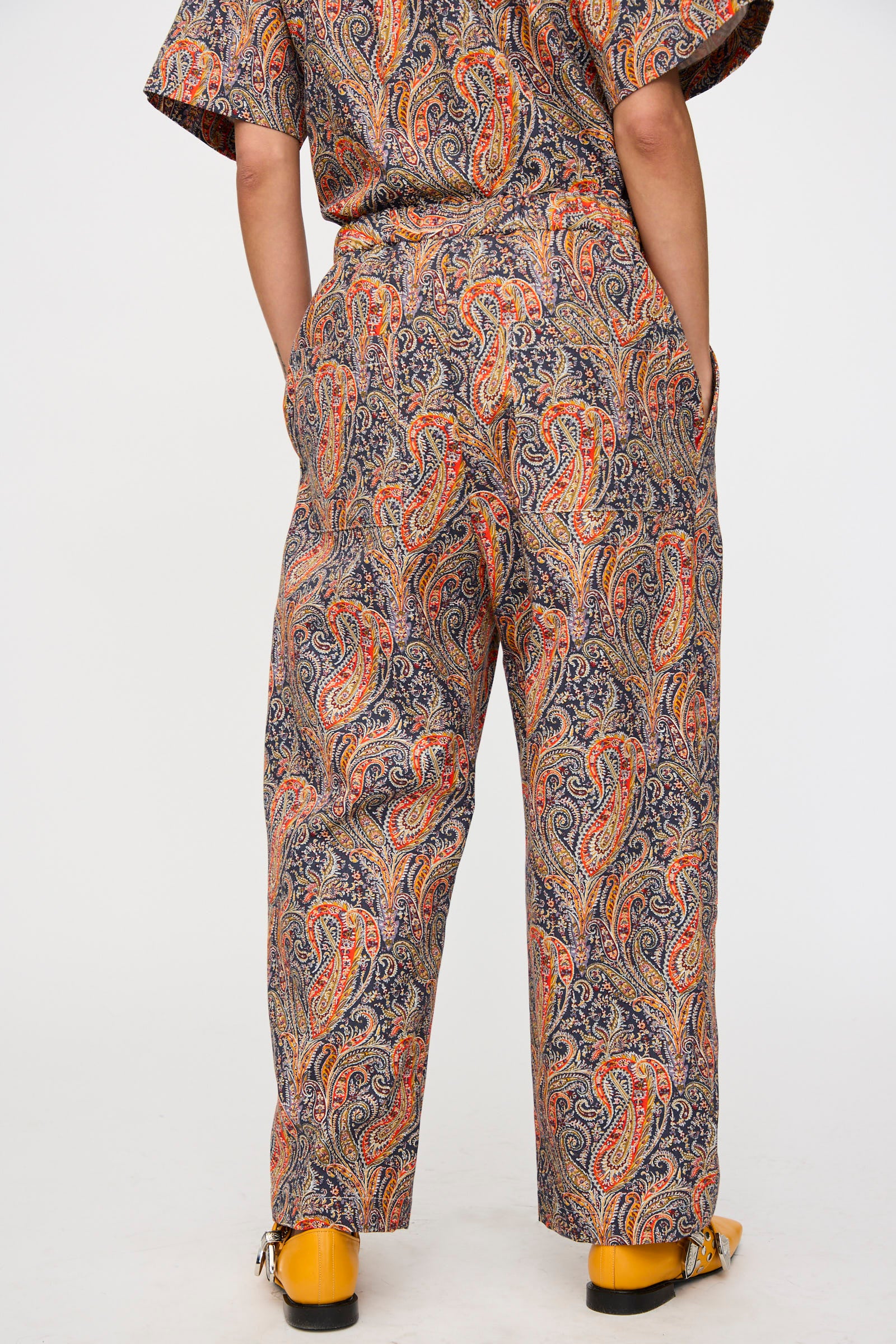 A person stands against a plain white background wearing the KasMaria Liberty Cotton Canvas Work Pant in Paisley with a matching cotton canvas shirt in orange, blue, and beige tones, paired with yellow shoes.