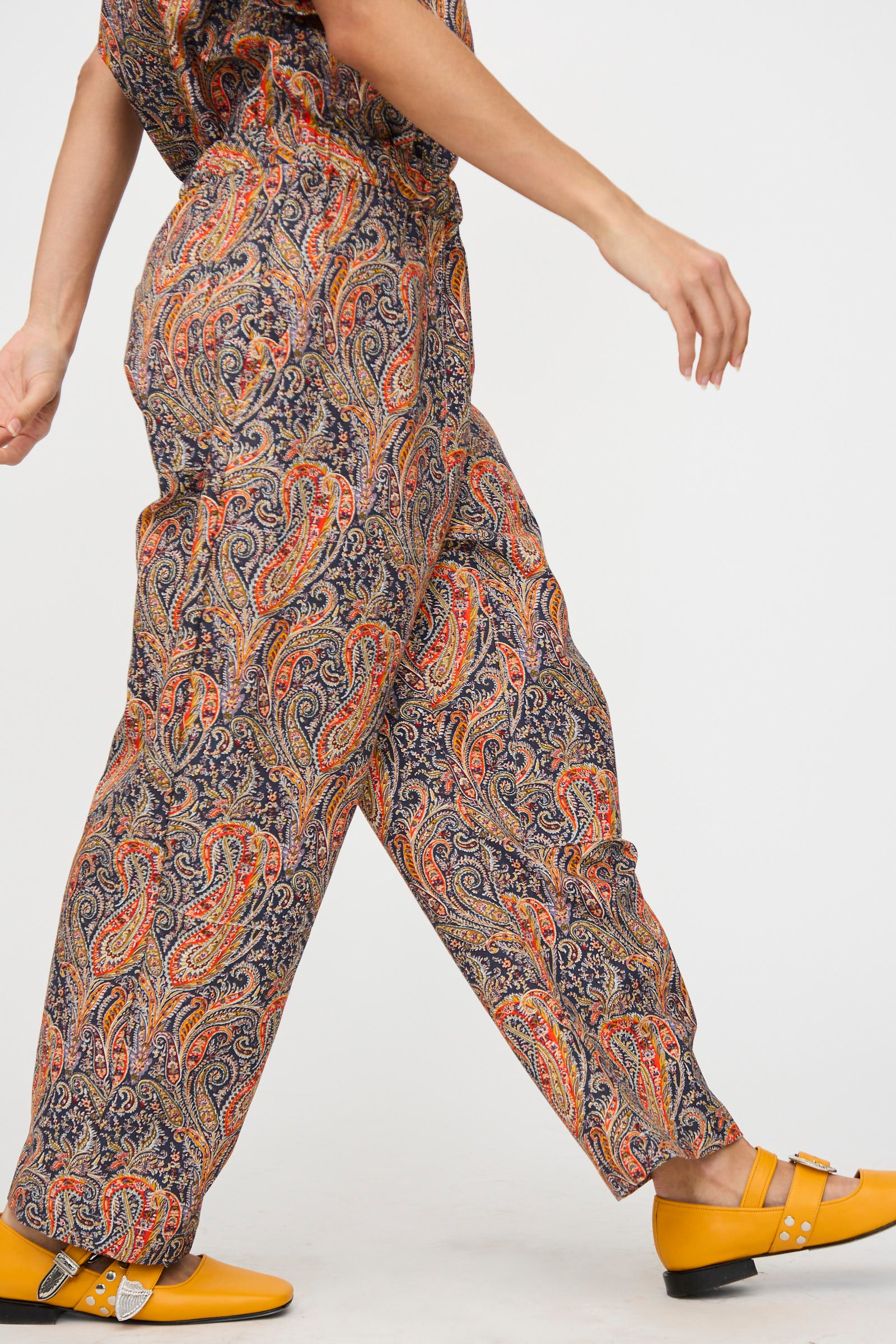 A person walks against a plain background, wearing KasMaria's Liberty Cotton Canvas Work Pant in Paisley—relaxed fit pants made in NYC with a colorful pattern—paired with bright yellow buckle shoes.