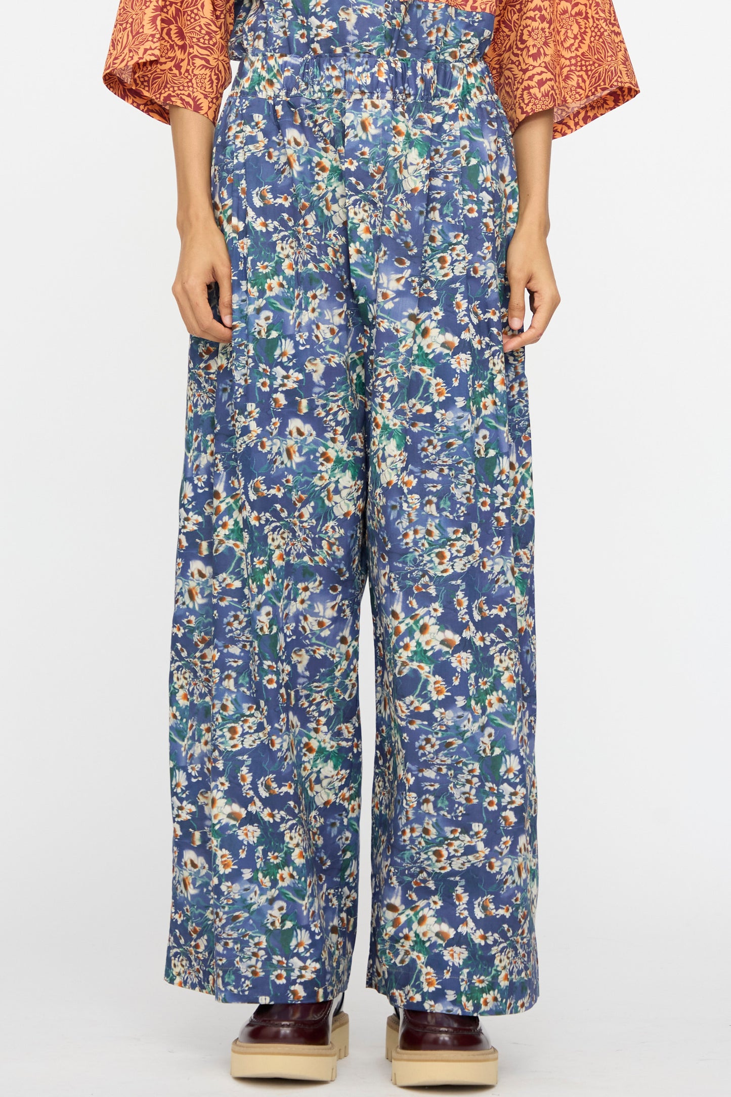 Model wears the KasMaria Liberty Floral Baggy Pleated Trouser in Blue, crafted in NYC from Liberty of London Tana Lawn Cotton, paired with a patterned orange top and brown platform shoes against a white background.