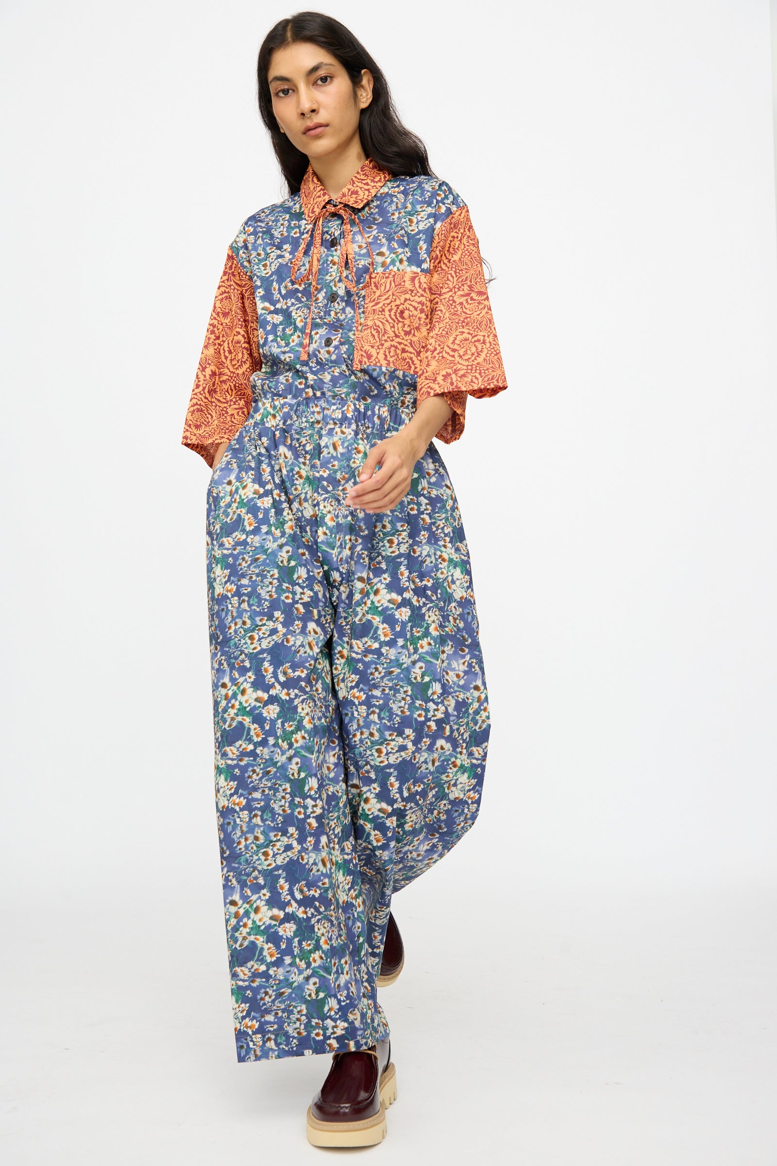 A woman walks in KasMaria's Liberty Floral Baggy Pleated Trouser in Blue, featuring orange patterned collar, pocket, and sleeves, styled with platform shoes.