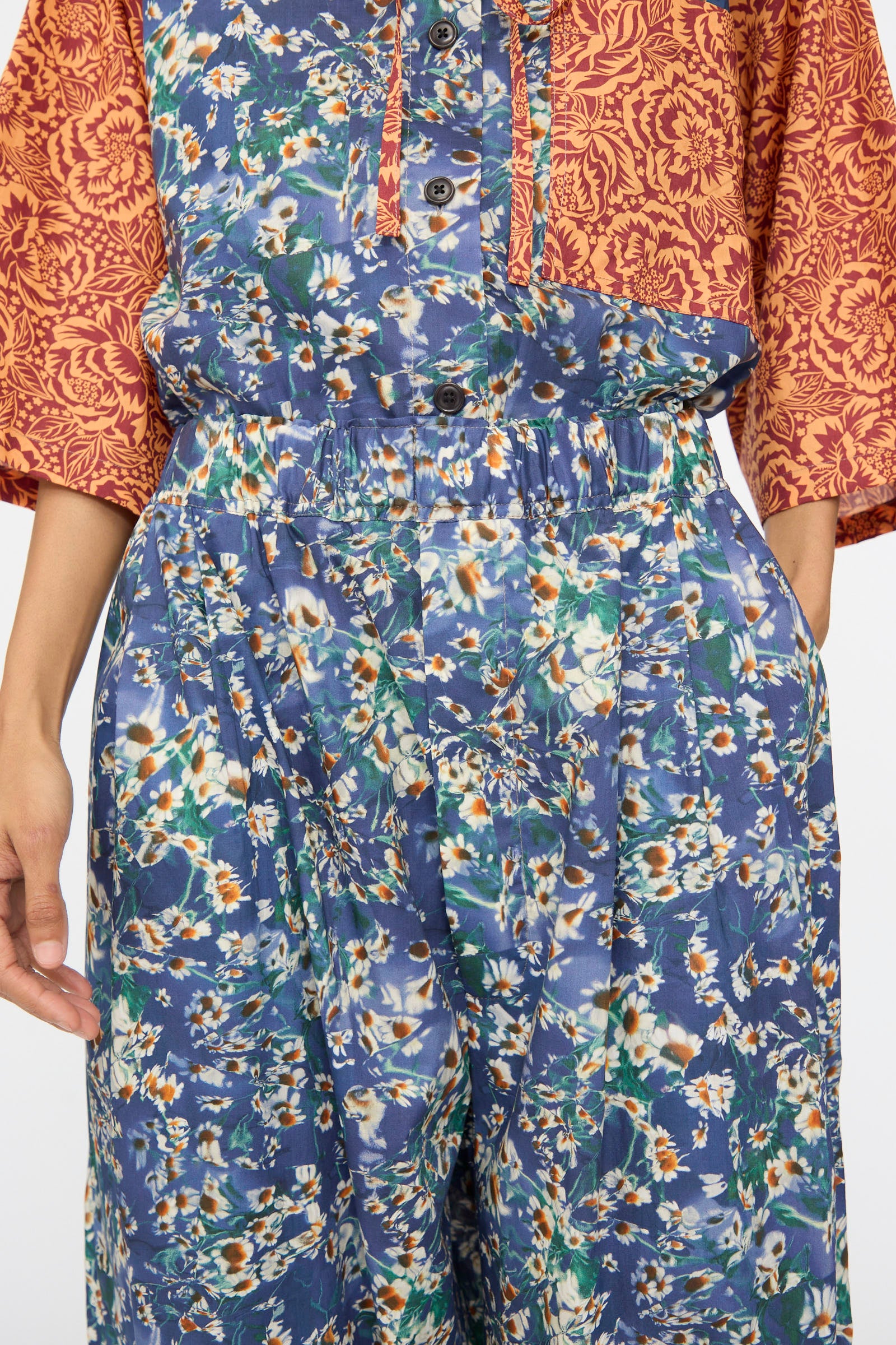 A person shown from shoulders to knees wears a red patterned top and KasMaria Liberty Floral Baggy Pleated Trouser in Blue, with hands in the pockets.